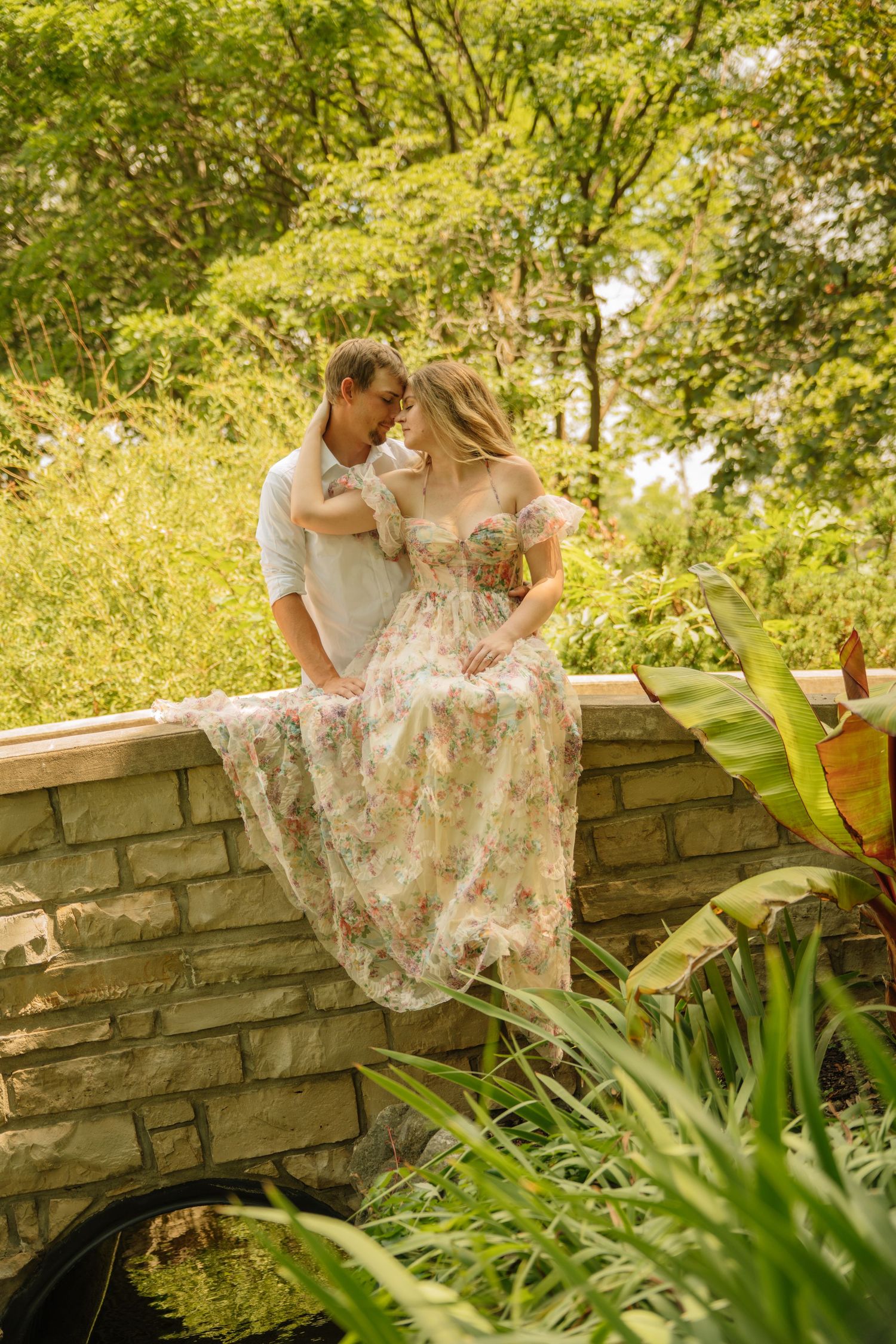 A woman in a floral dress sits on a stone bridge wall with her partner in a sunlit garden setting.