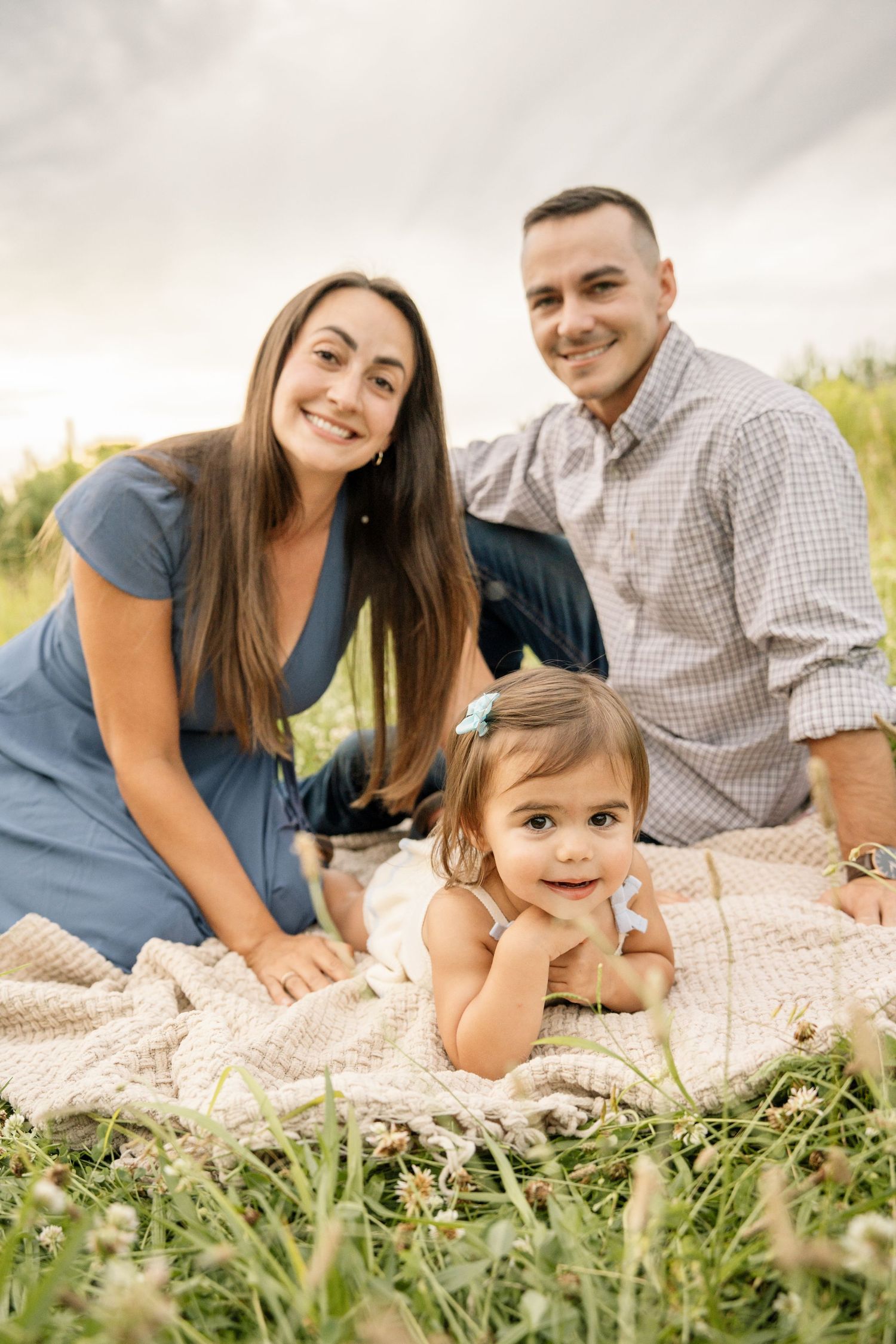 Happy young family with toddler daughter relaxing on blanket in grassy field.