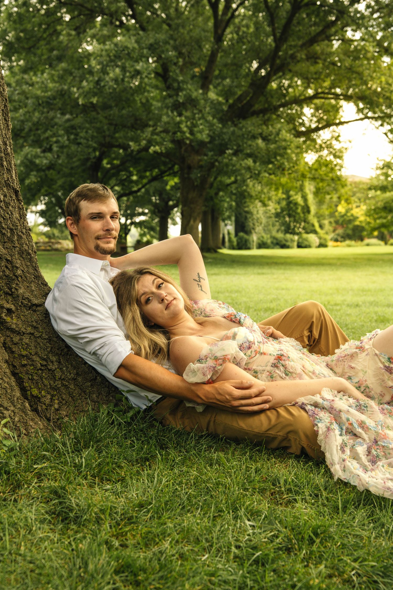 Couple relaxing against a tree trunk on green grass, woman in floral dress lying in mans arms outdoors.