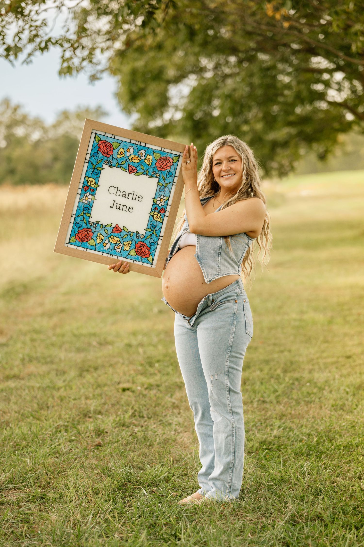 Glowing pregnant woman smiles holding a colorful framed Charlie June name sign outdoors in a park.