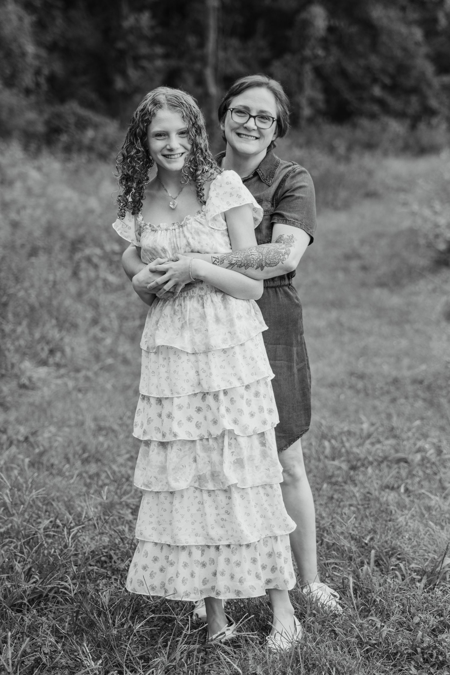 Black and white photo of a mother hugging her curly-haired daughter in a ruffled dress outdoors.