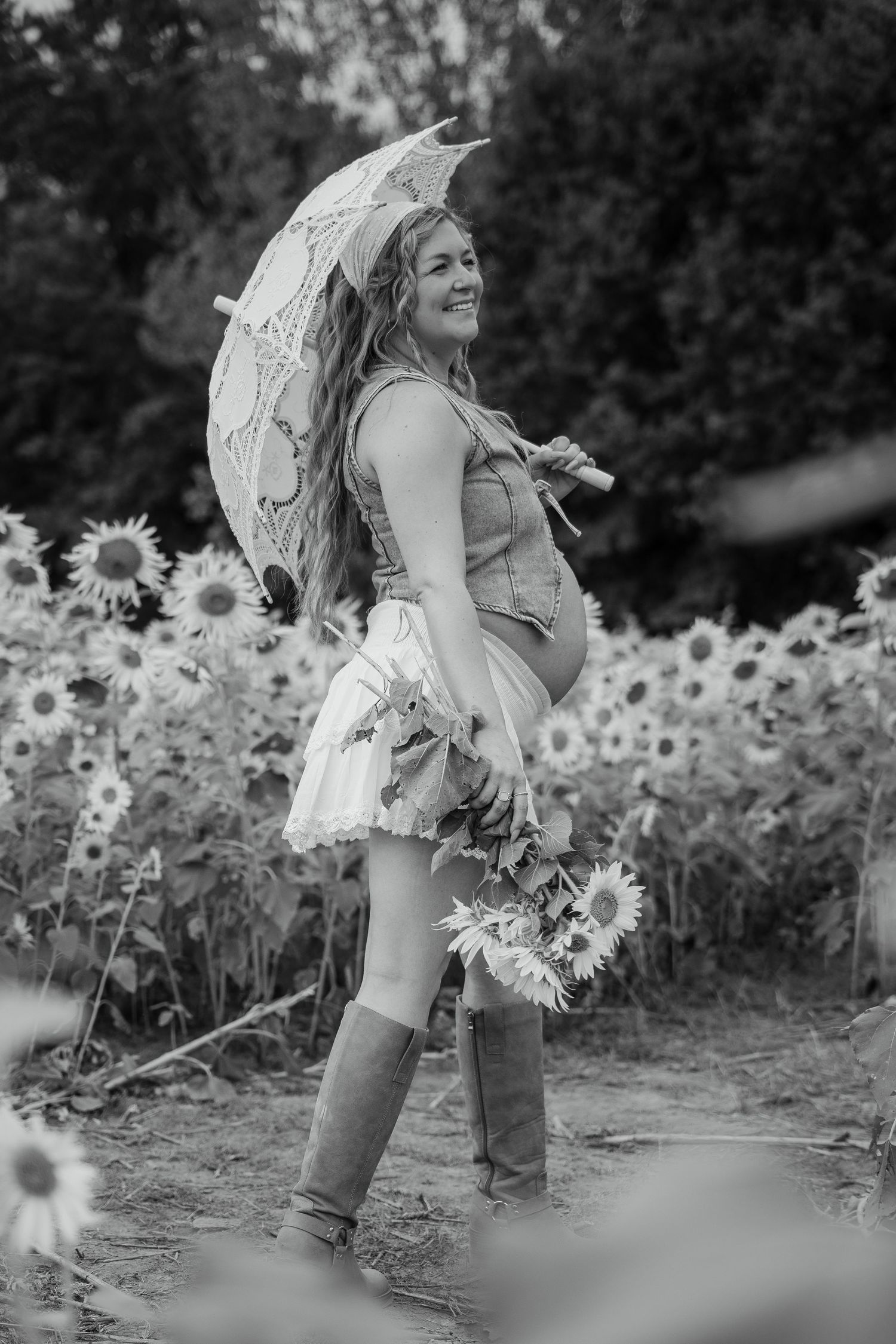 Pregnant woman in sunflower field holding lace umbrella and sunflowers, wearing skirt and boots, black and white photo.