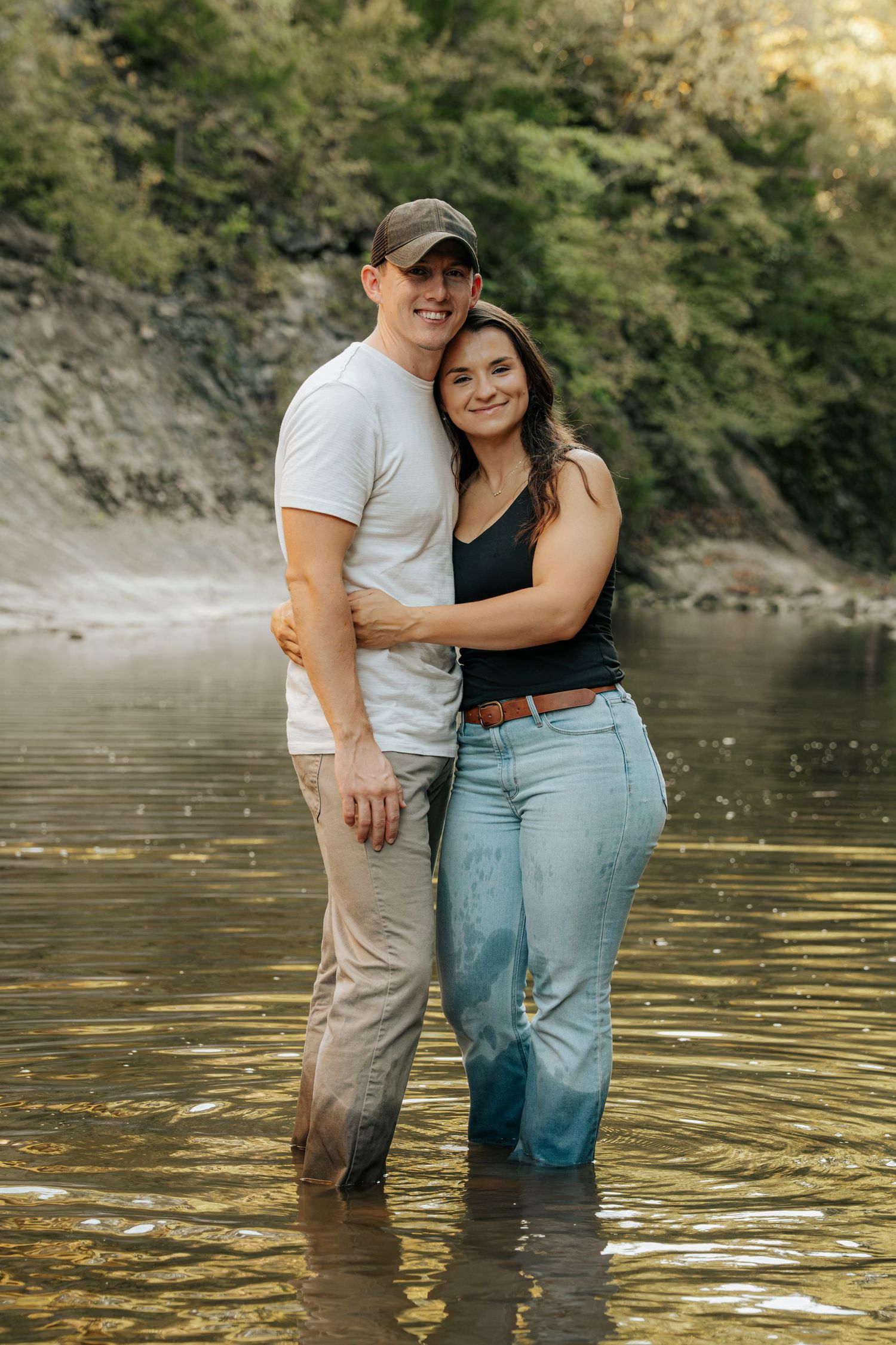 A couple embracing and smiling while standing in a shallow river, surrounded by lush green trees.