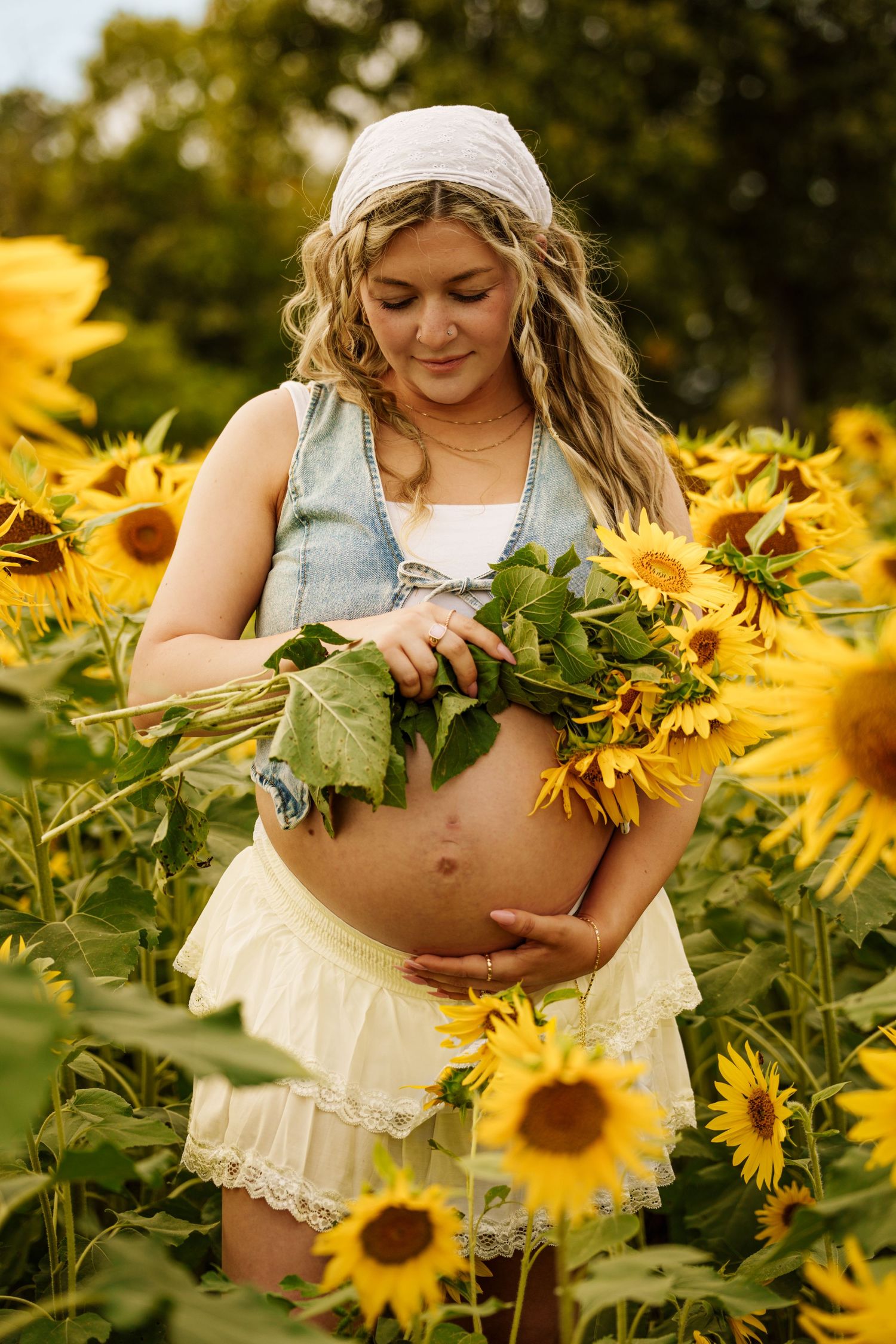 Smiling pregnant woman cradles sunflowers and baby bump in a bright yellow sunflower field.