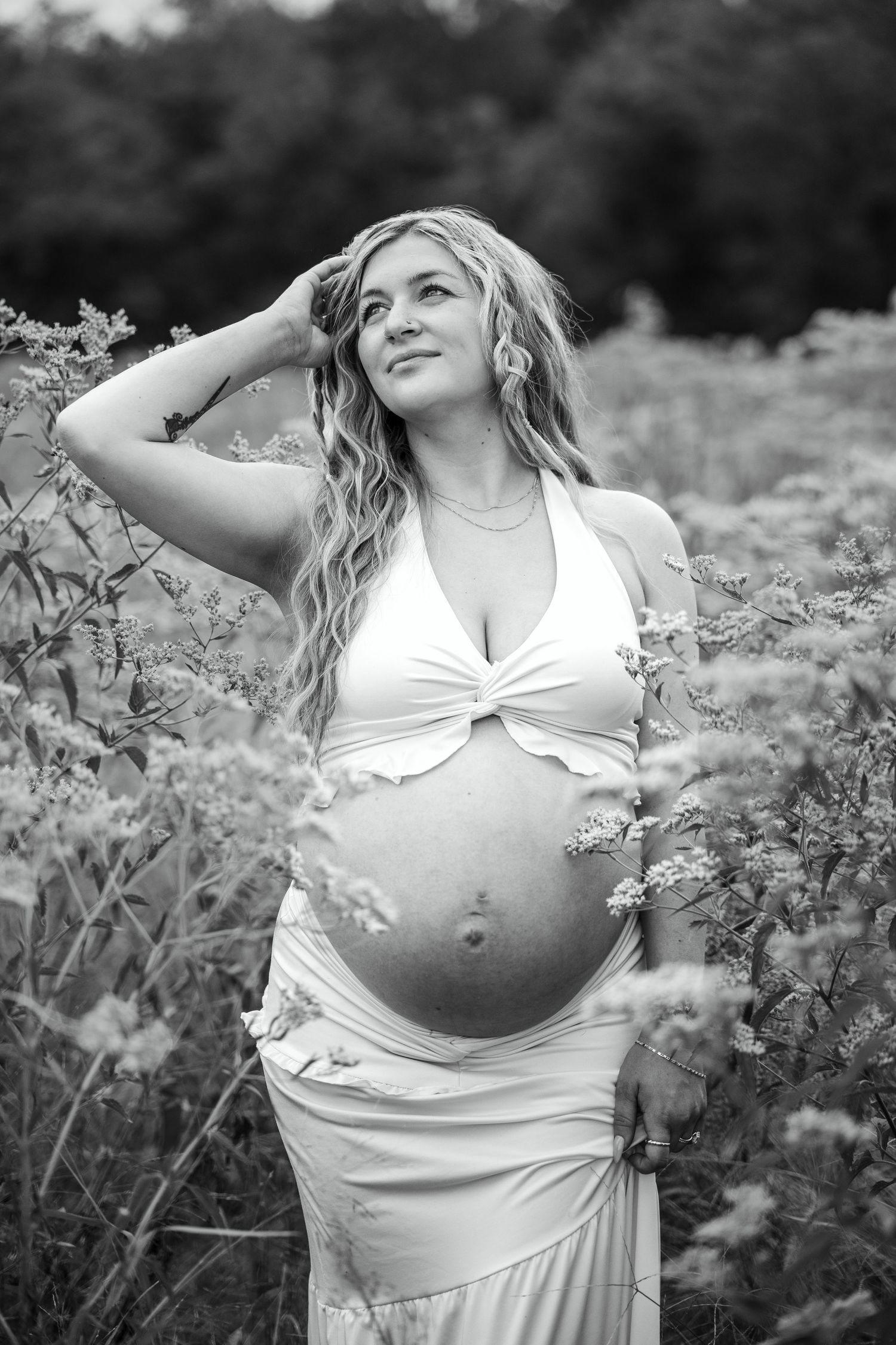 Black and white photo of pregnant woman in white outfit posing in wildflower meadow.