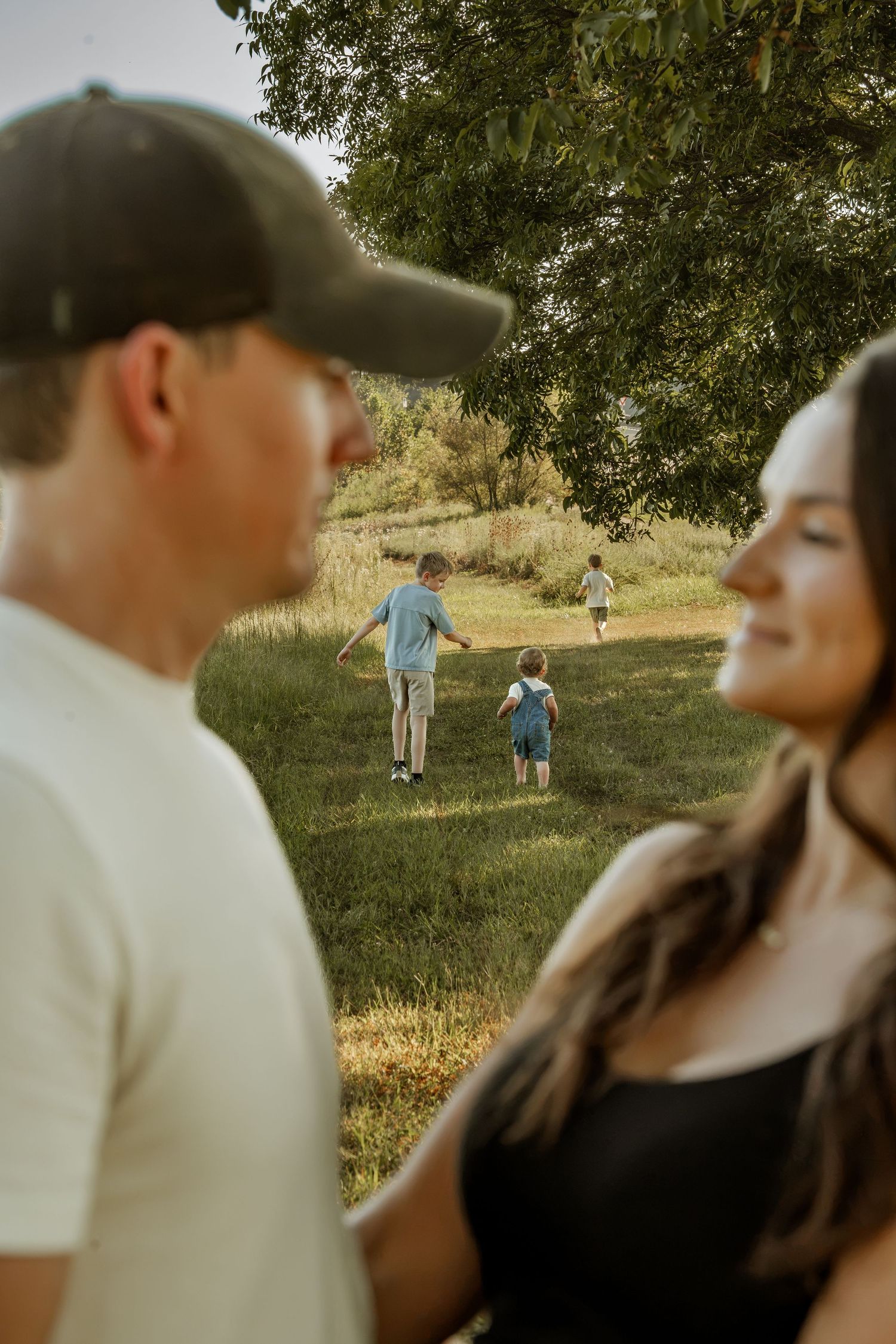 Parents stand in foreground while three children play in green field under large tree during family outdoor photo session.
