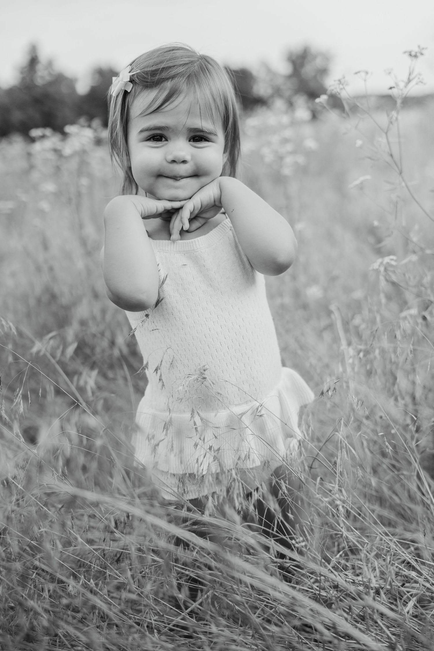 Black and white portrait of a cute toddler girl in a white dress posing in a meadow.