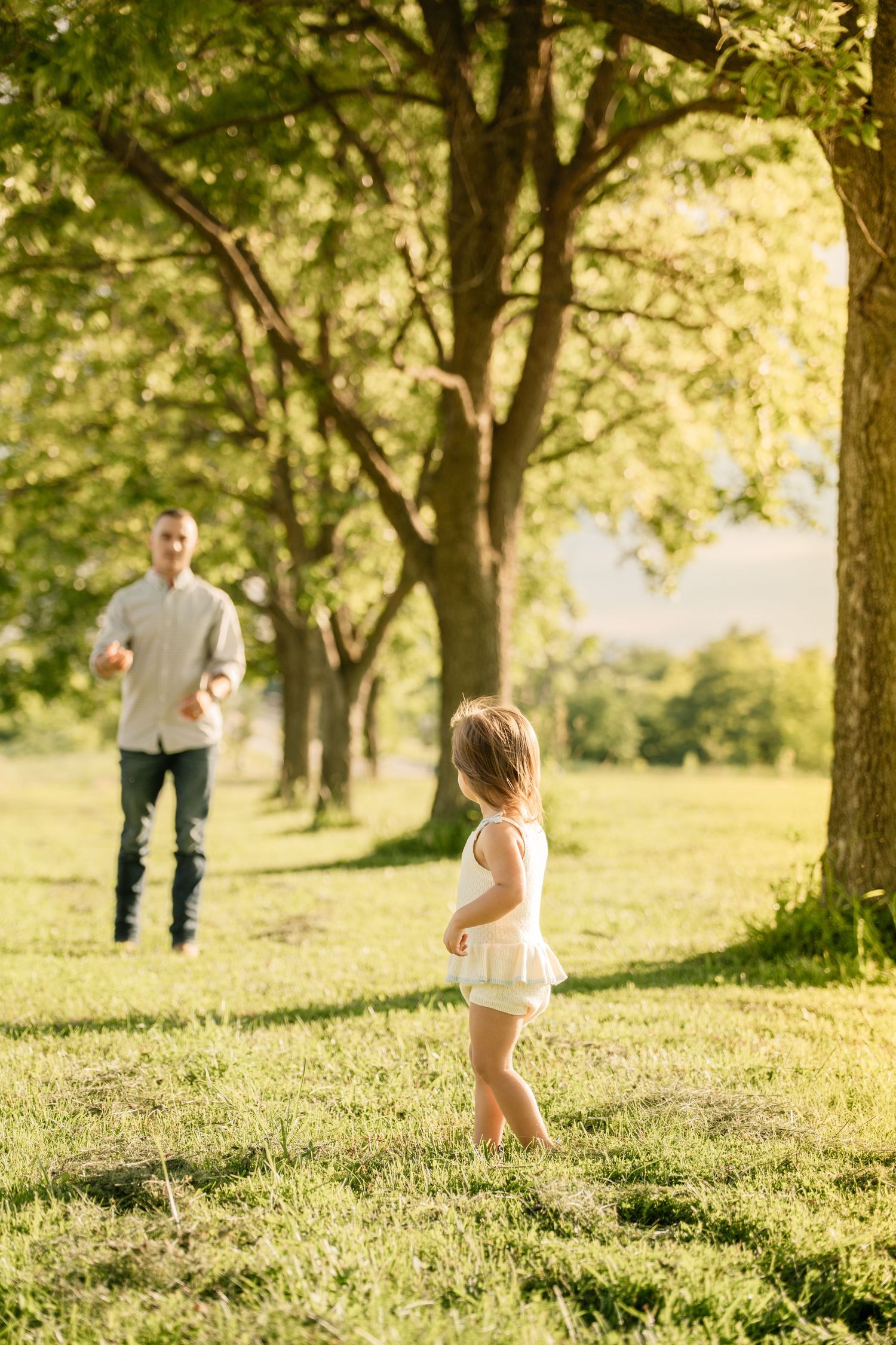 Father and young daughter playing catch together in a sunny green park with tall trees.