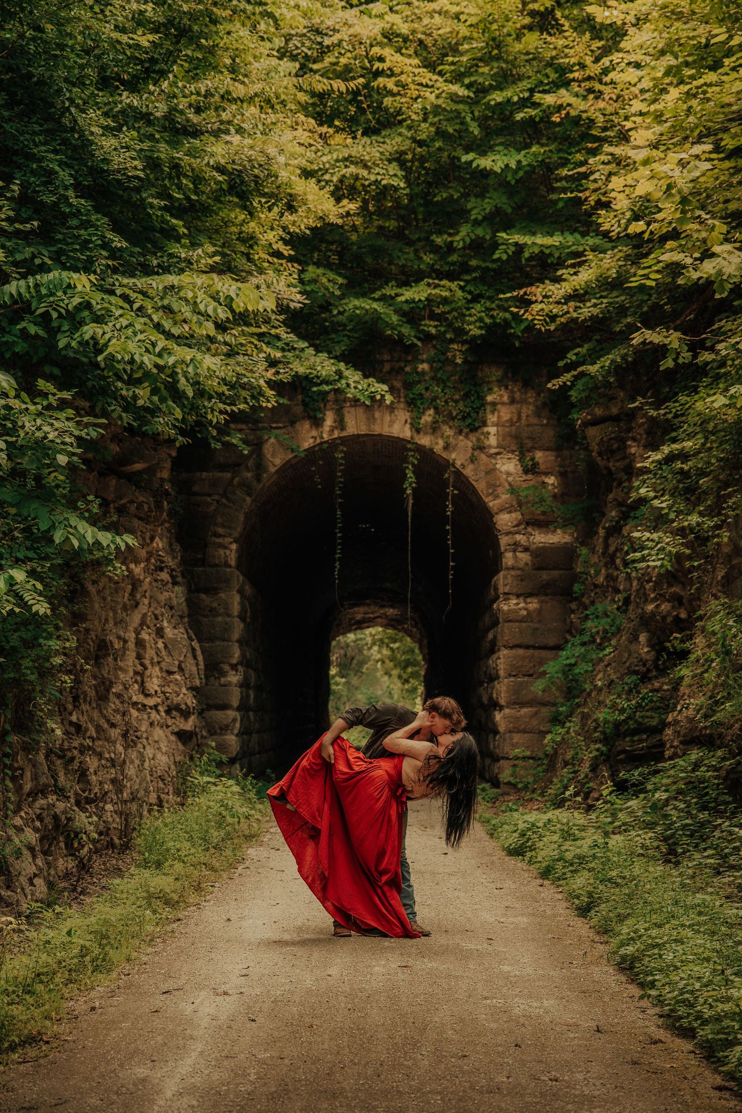 Couple dancing in red dress before stone tunnel arch surrounded by lush green forest trees on a dirt path.