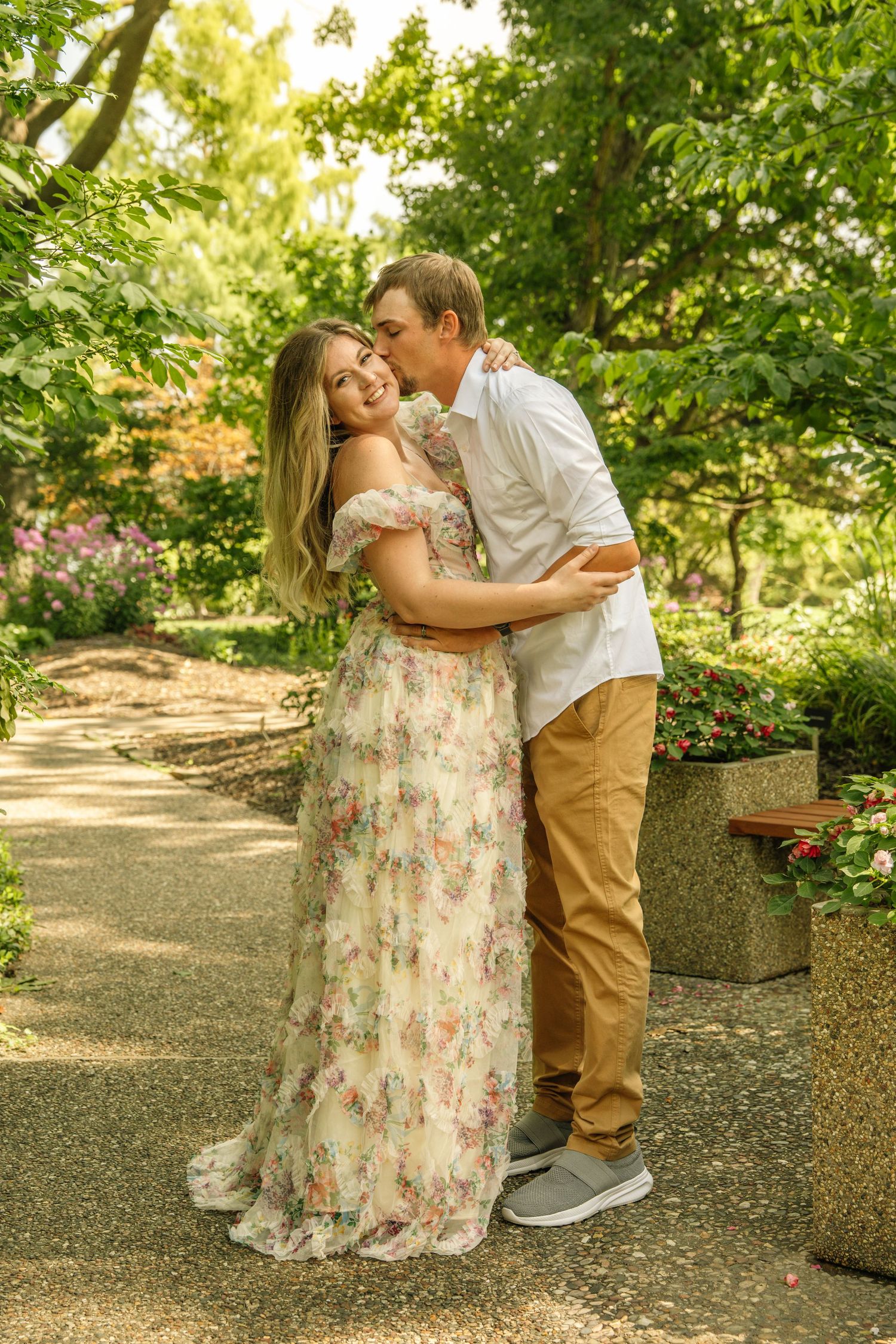 Man in white shirt kissing woman in floral dress on the cheek along a sunny garden pathway.