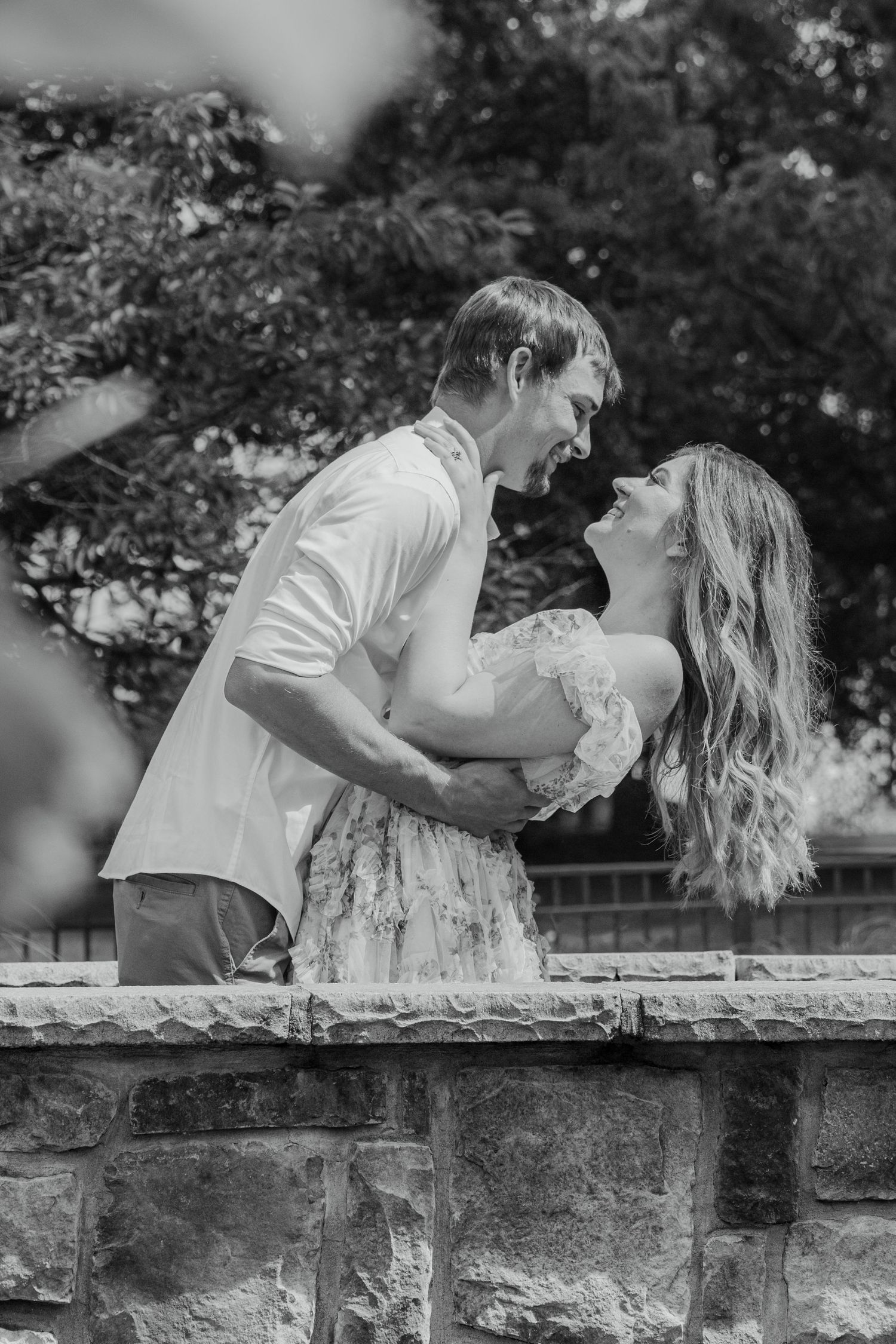 Black and white photo of a couple embracing and laughing together outdoors near a stone wall with trees behind them.