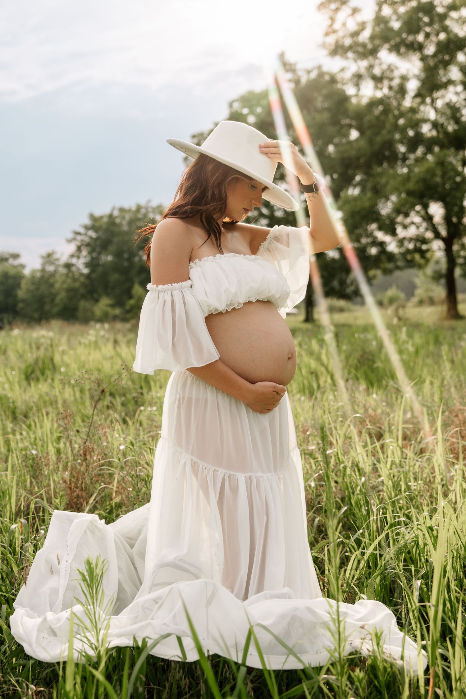 Pregnant woman in white off-shoulder dress and hat holding her belly in a sunlit meadow.