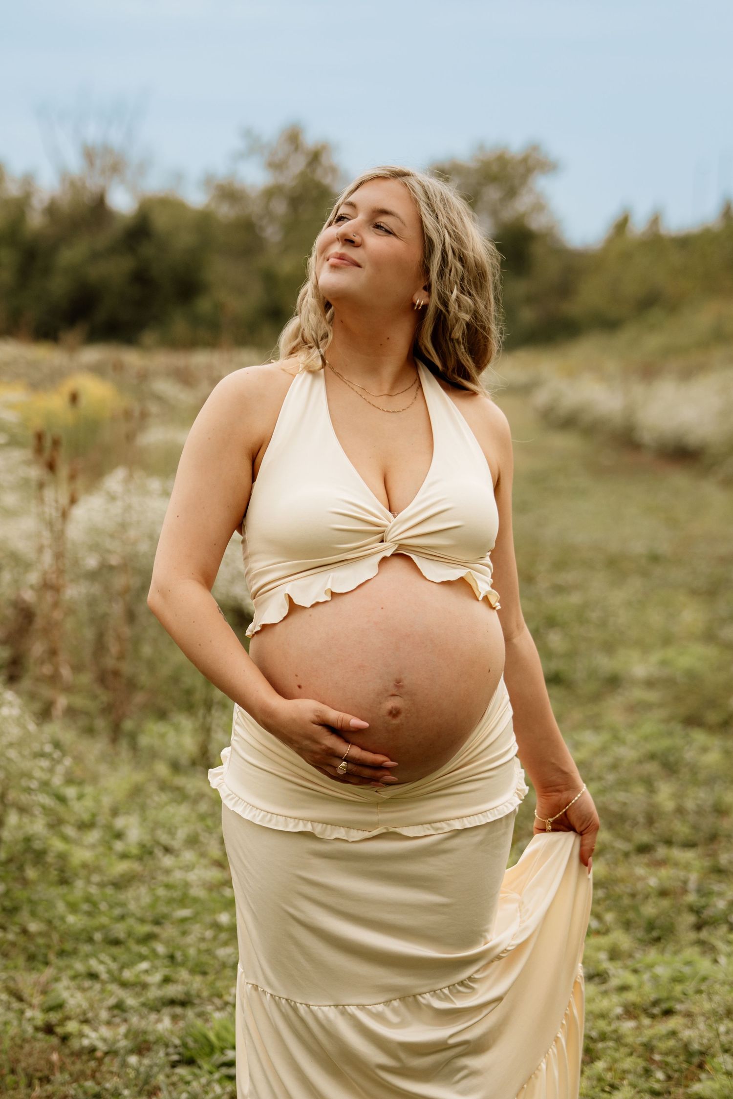 Pregnant woman in cream halter crop top and skirt cradling baby bump outdoors in a natural field setting.