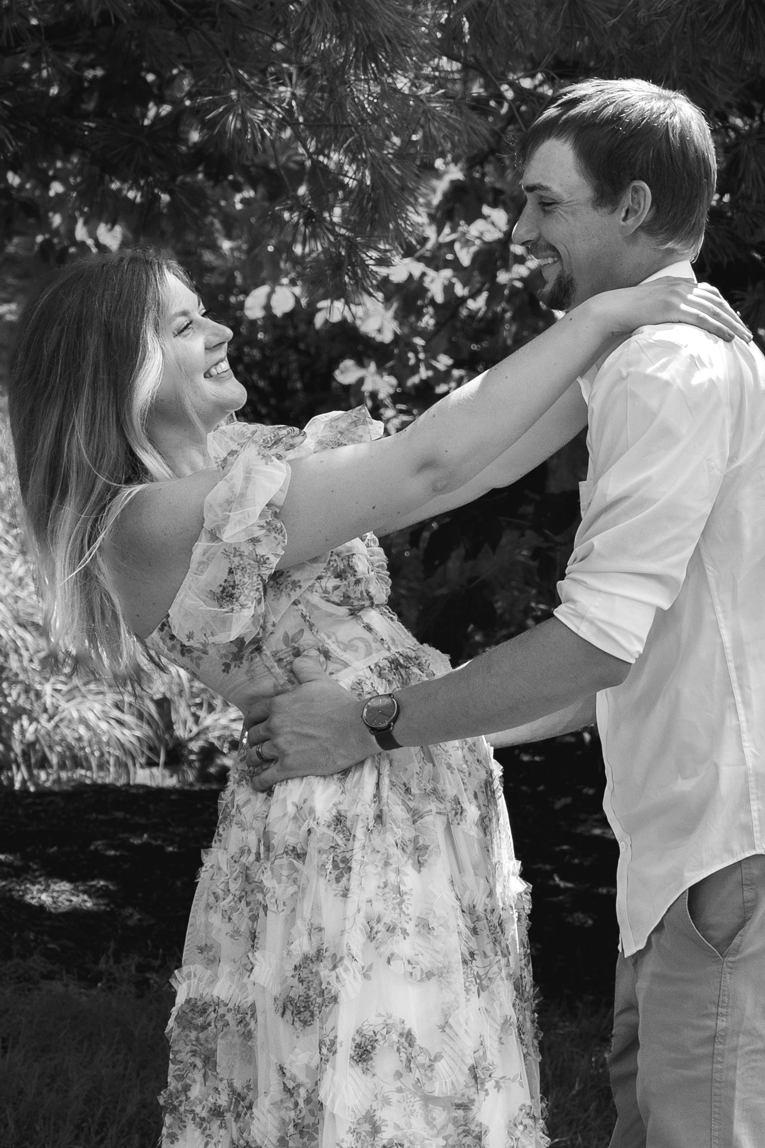 Black and white photo of a laughing couple embracing outdoors, woman in floral dress with hands on mans shoulders.