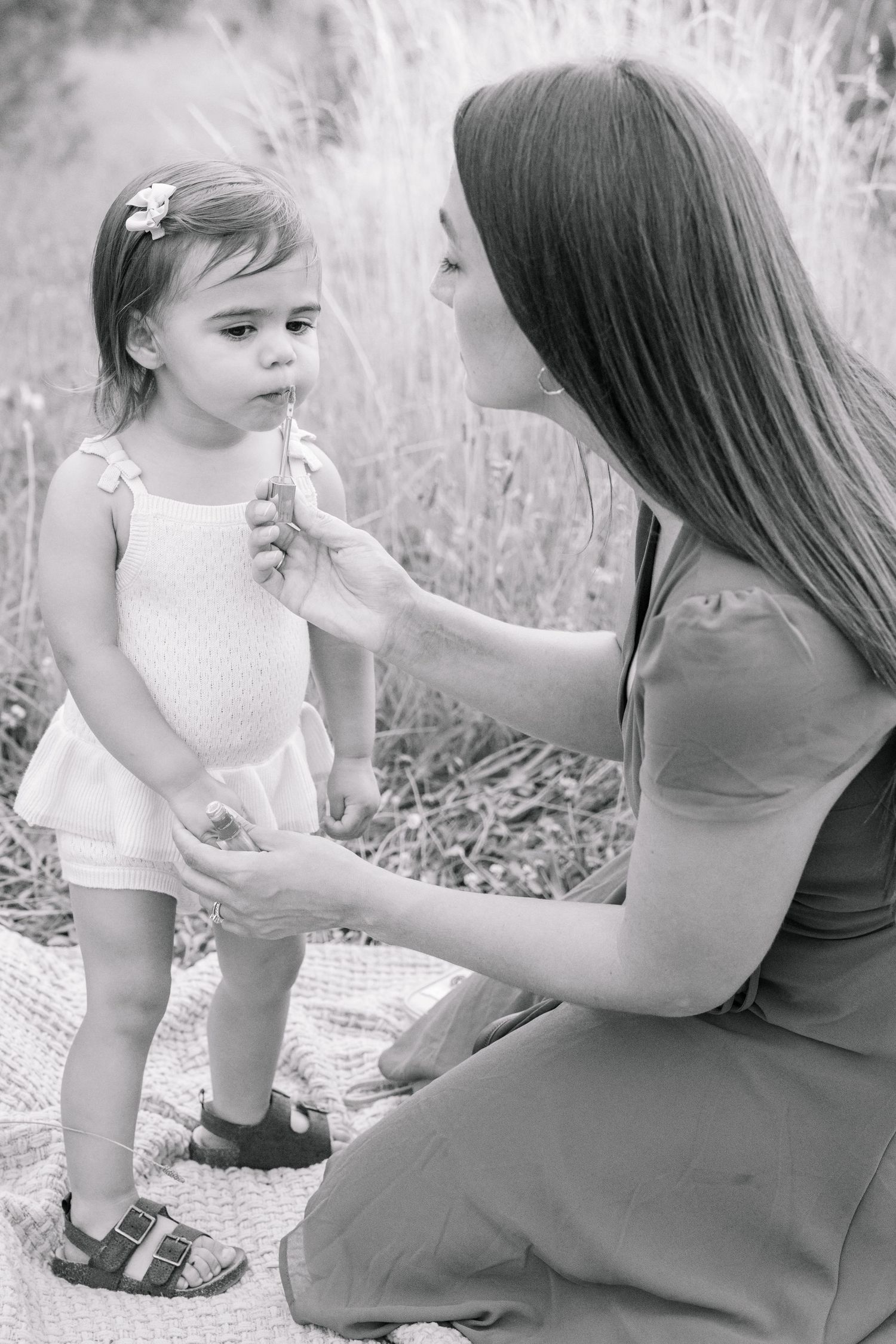 Black and white photo of mother offering flower to toddler daughter on blanket outdoors.