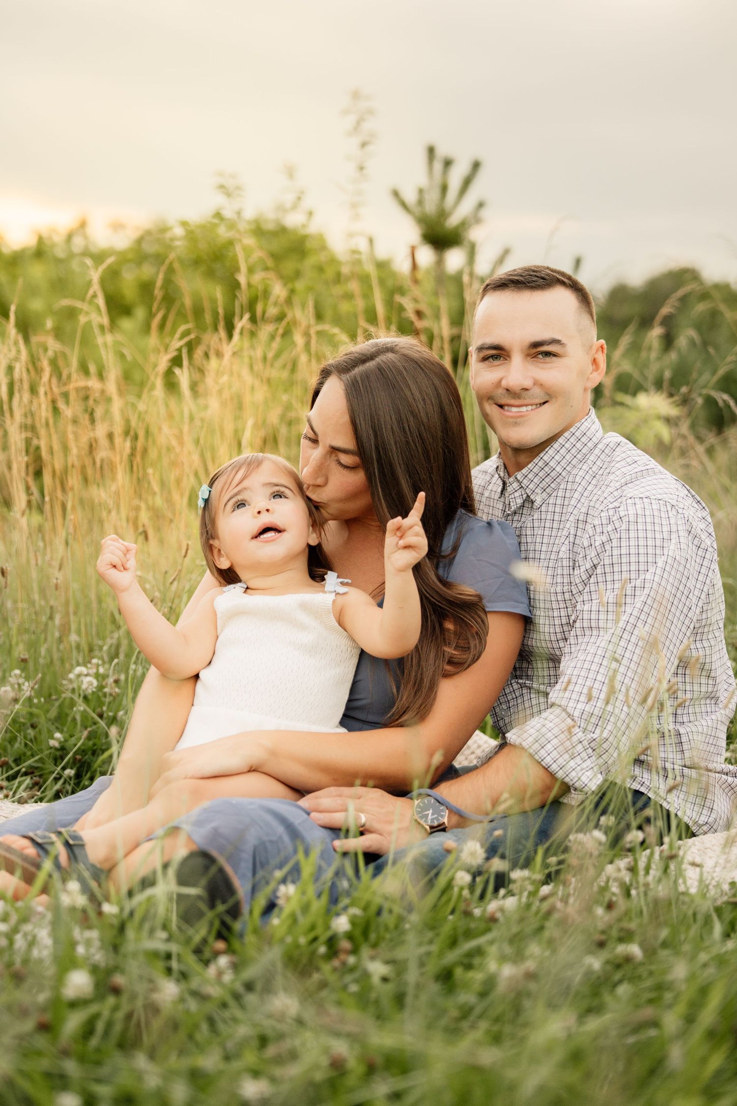 Happy young family sitting together in tall grass field at golden hour sunset.