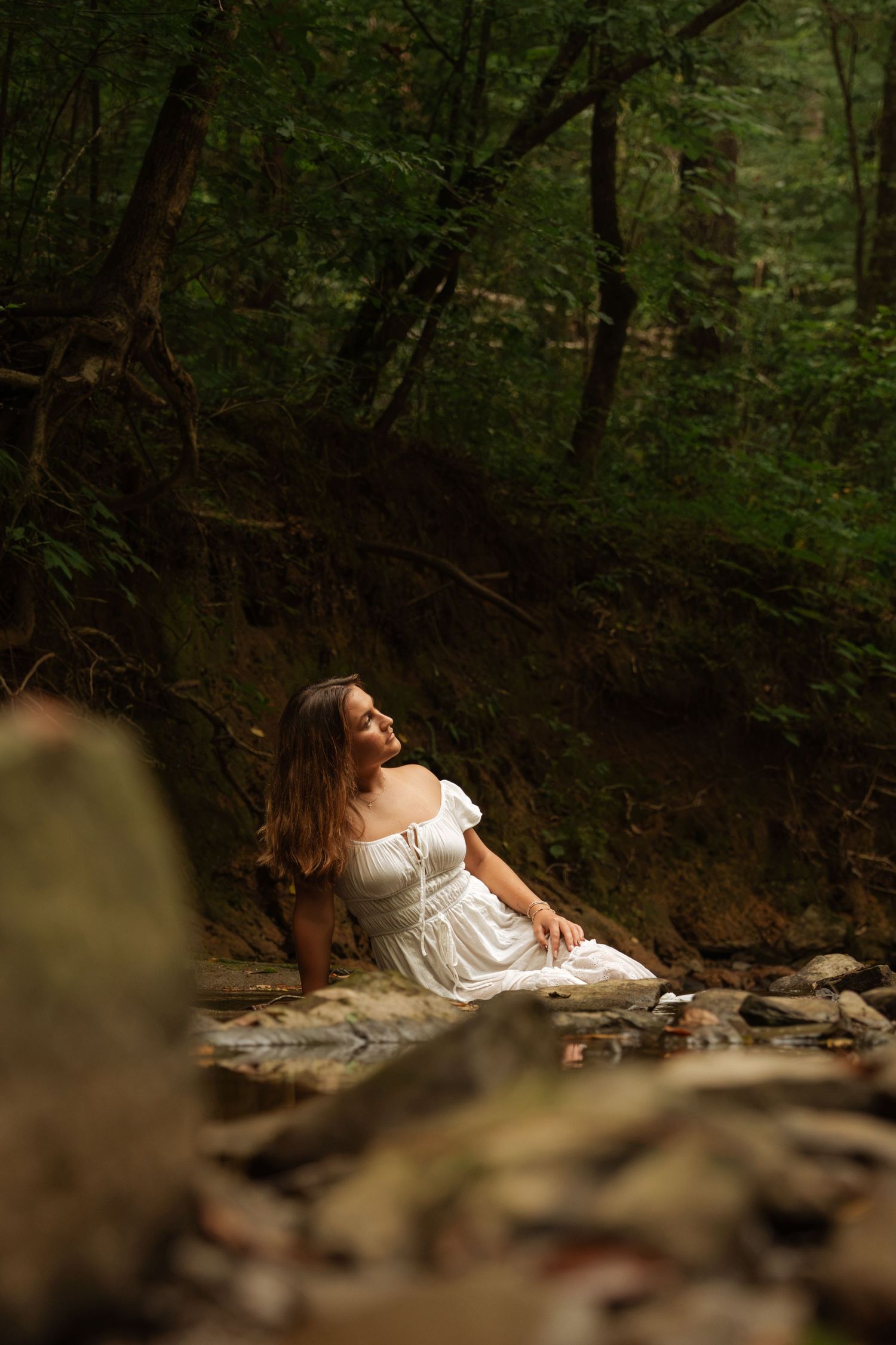 Young woman in white dress sitting peacefully on rocks in a lush green forest setting.