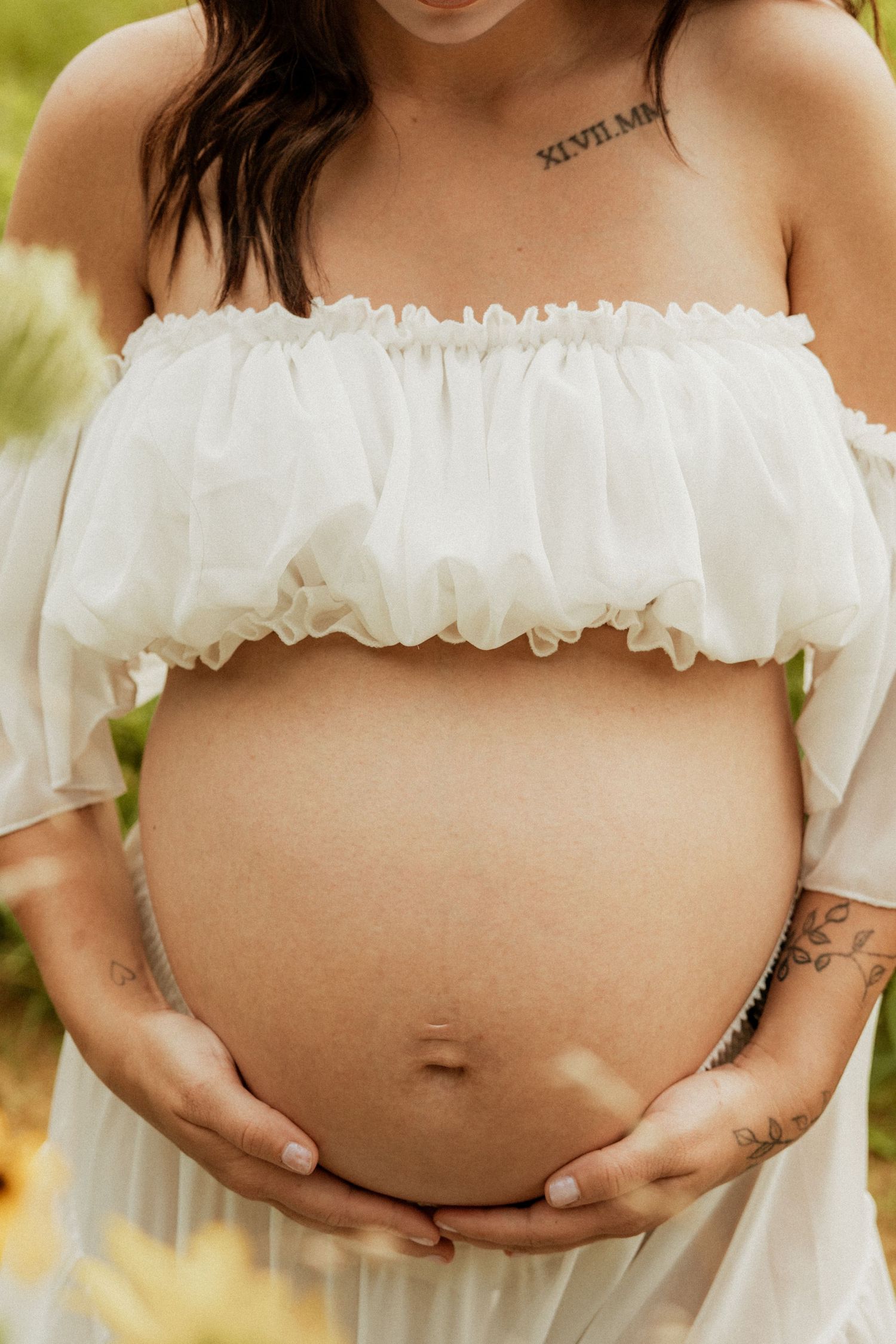 Close-up of a pregnant woman in a white ruffled top gently cradling her bare baby bump outdoors.