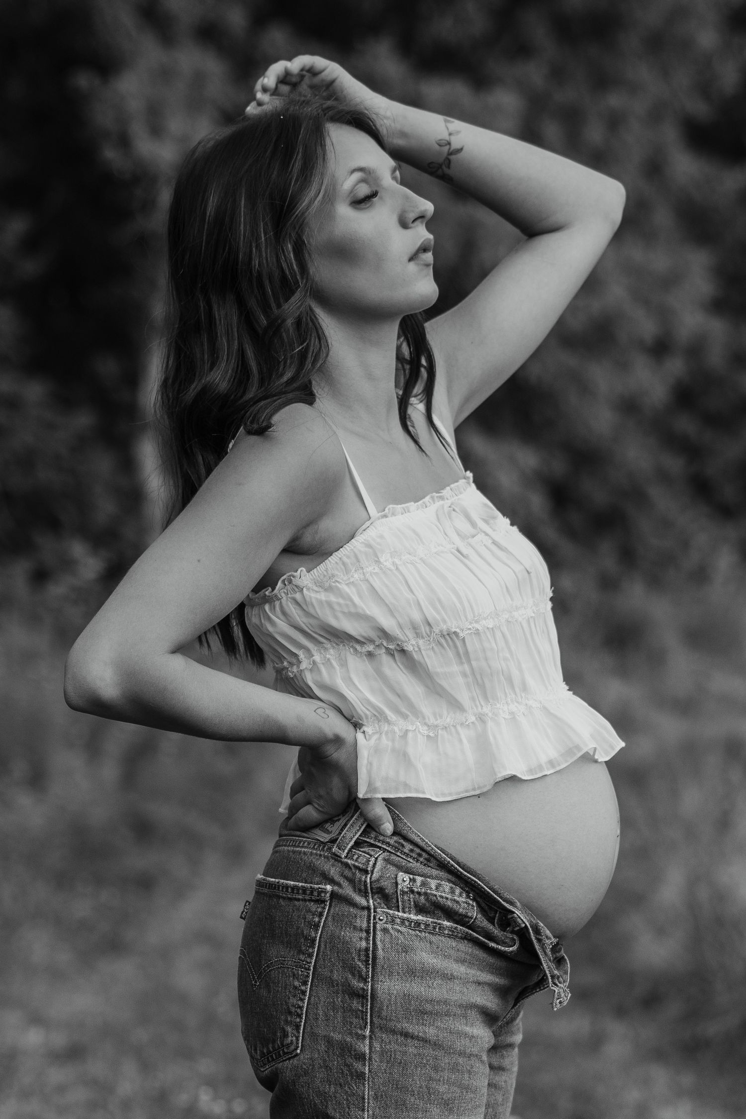 Black and white photo of a pregnant woman in a white crop top and jeans posing outdoors.