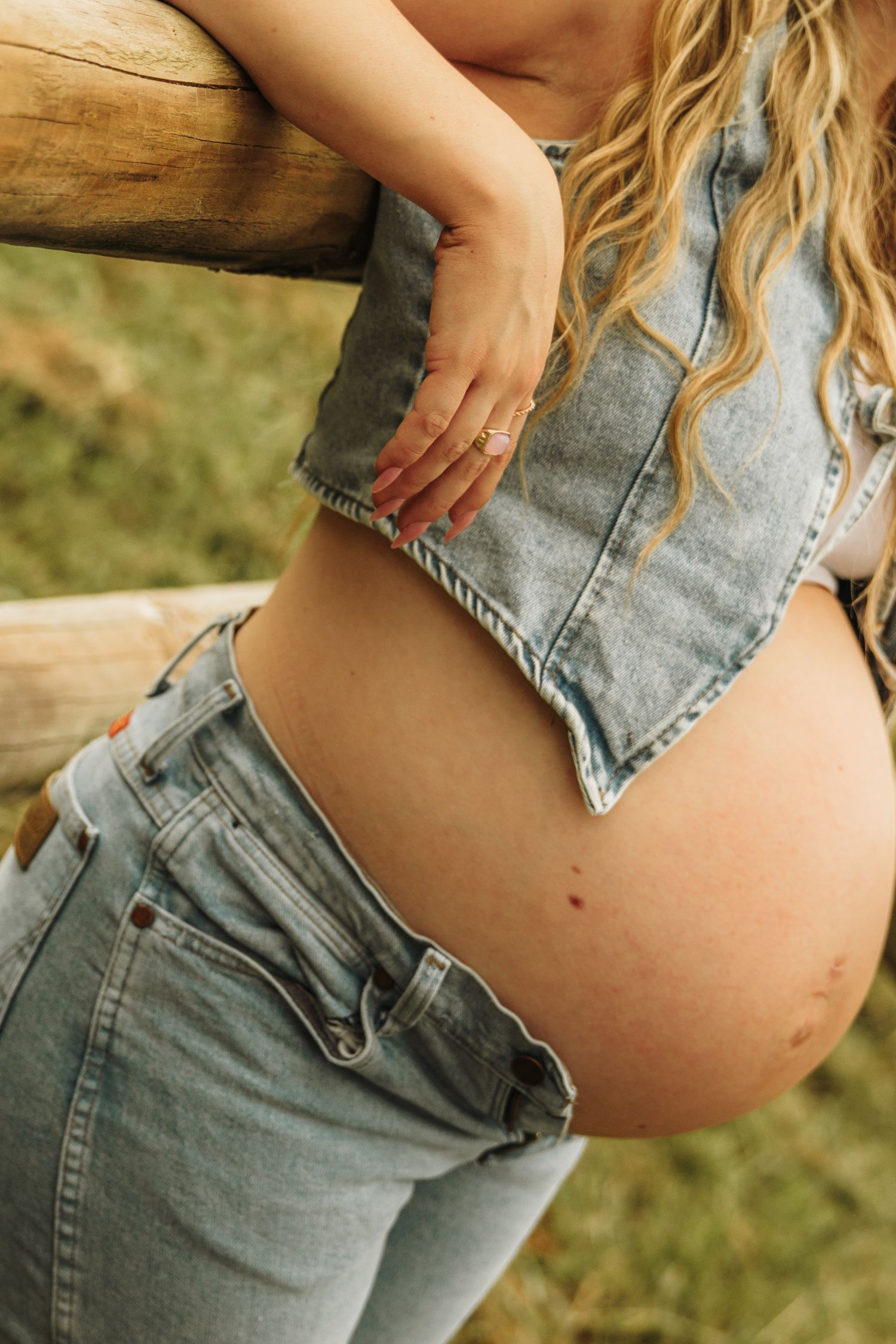 Pregnant woman in denim crop top and jeans showing baby bump outdoors near wooden fence.