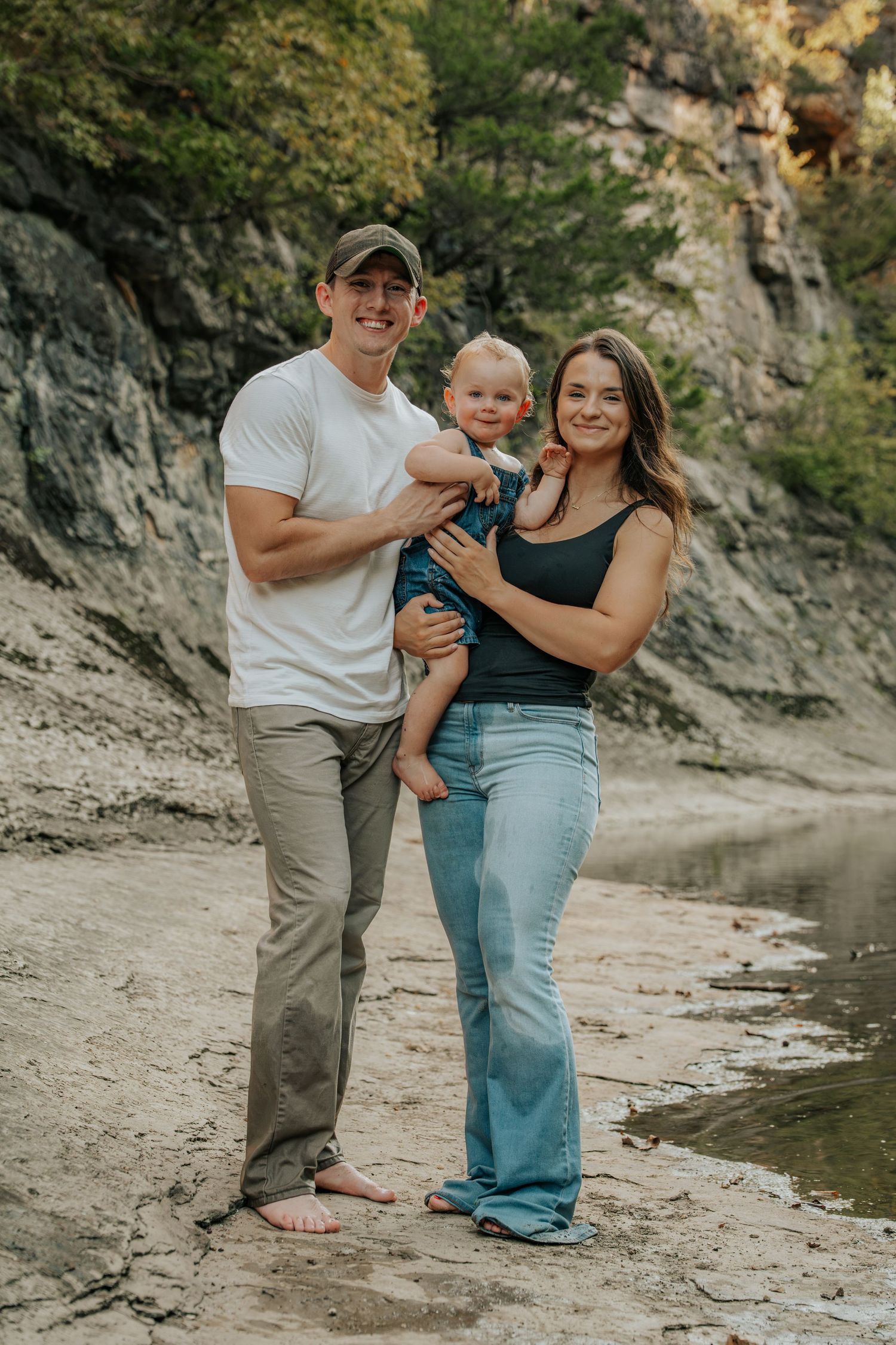 Happy young family with toddler posing on sandy riverbank near rocky cliffs.