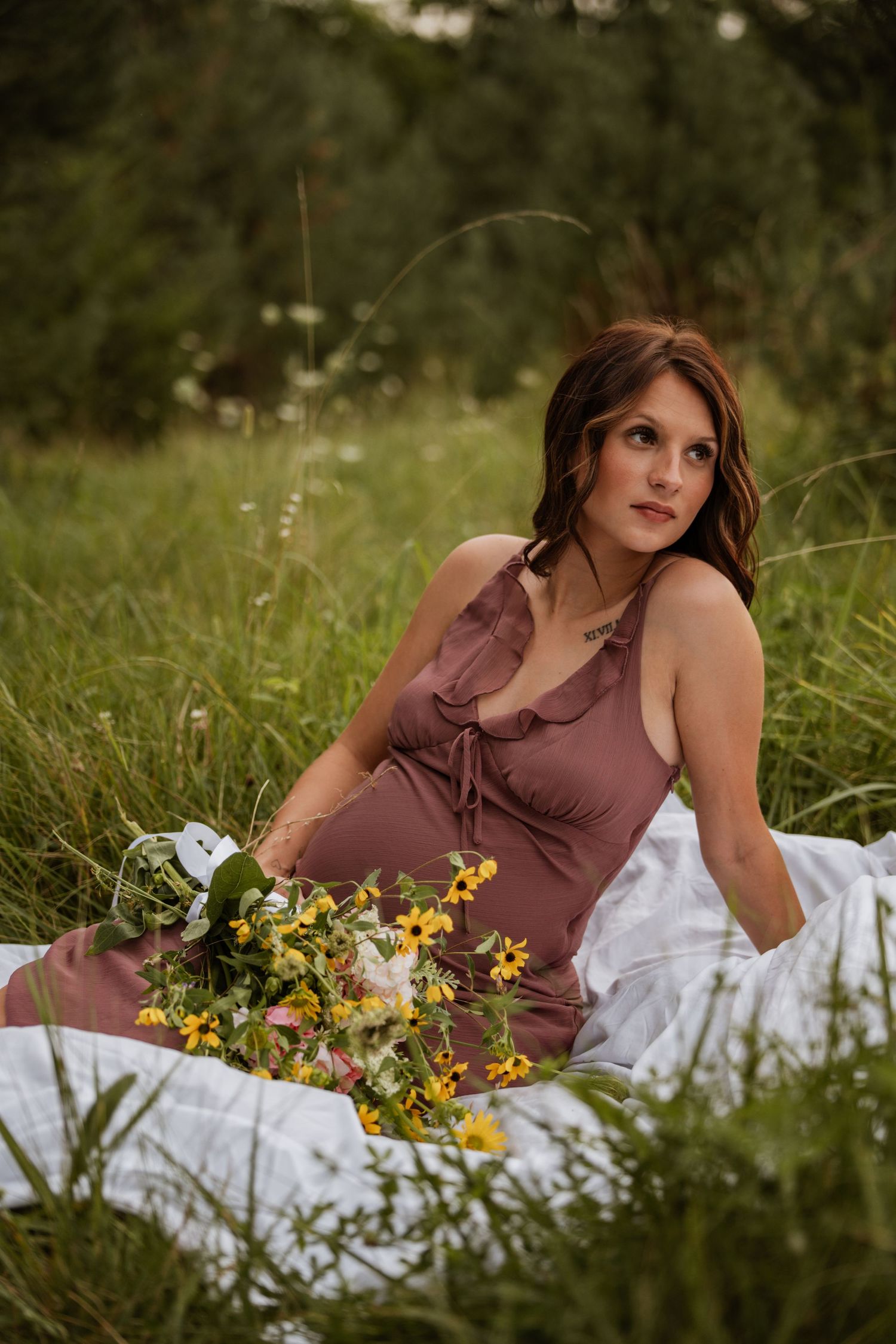 Pregnant woman in dusty rose slip dress reclines on white blanket in meadow with yellow wildflower bouquet.