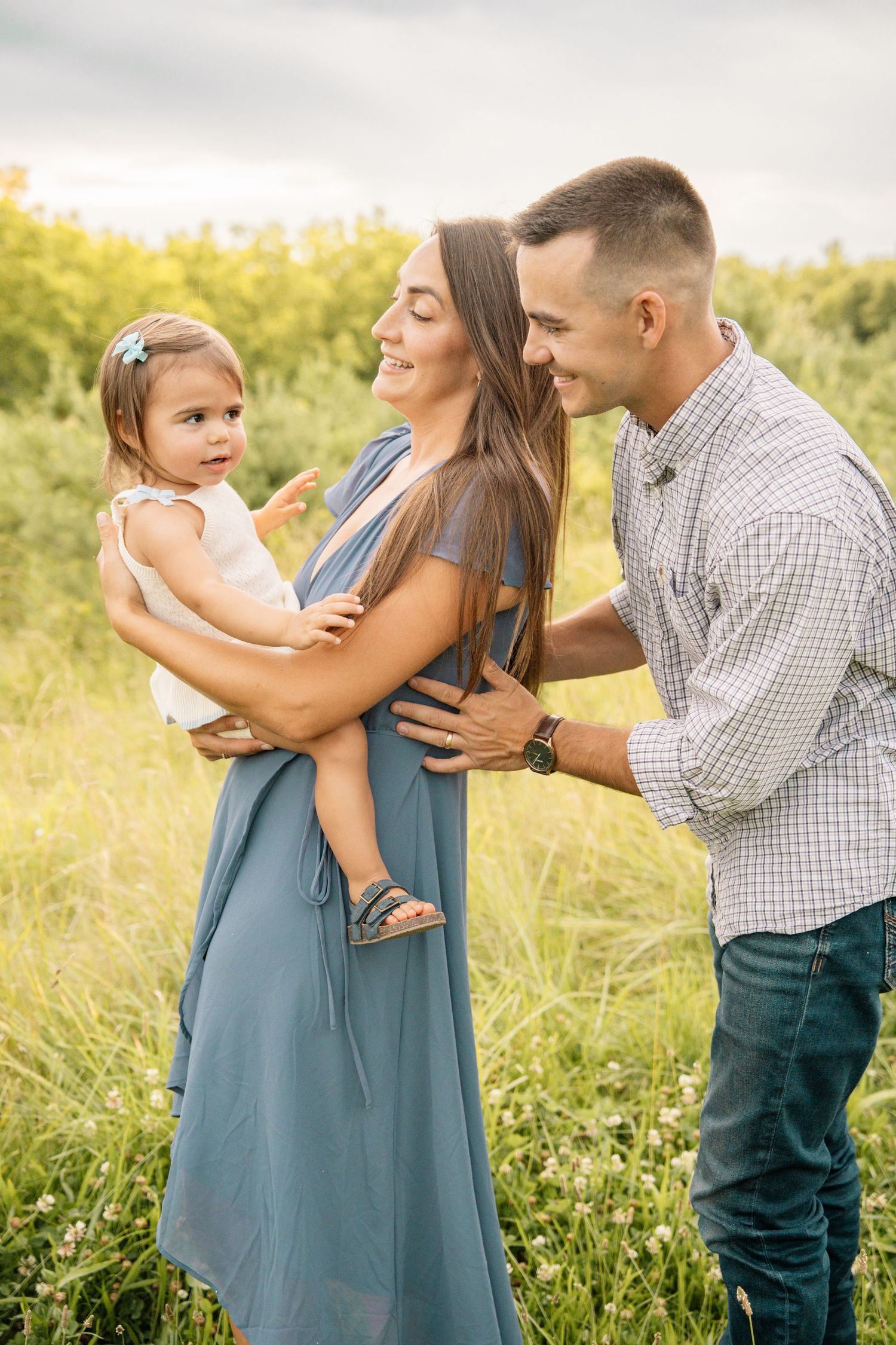 Young family with mother in blue dress and father holding toddler in green field.