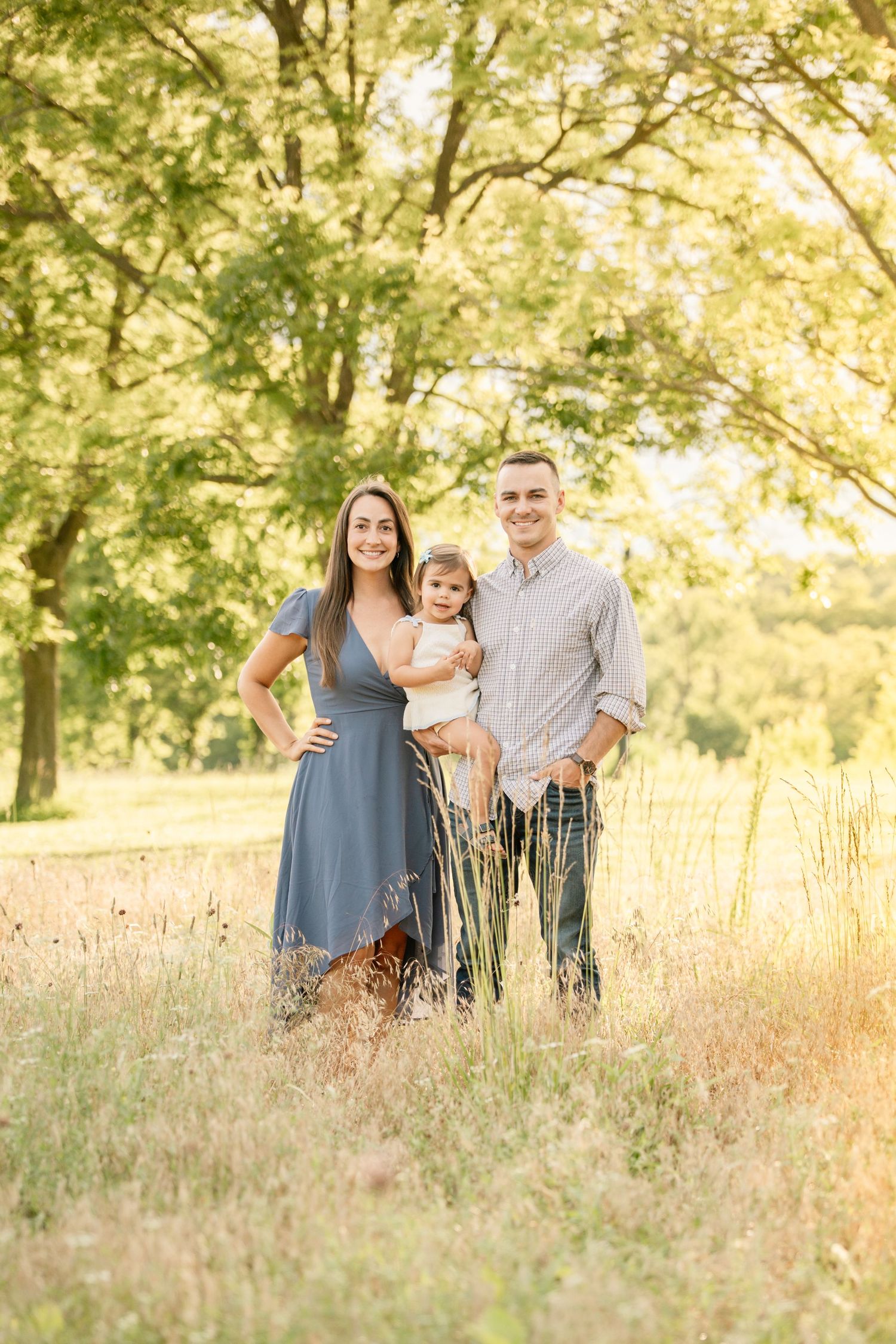 Young family of three posing together in a sunlit meadow surrounded by lush green trees.
