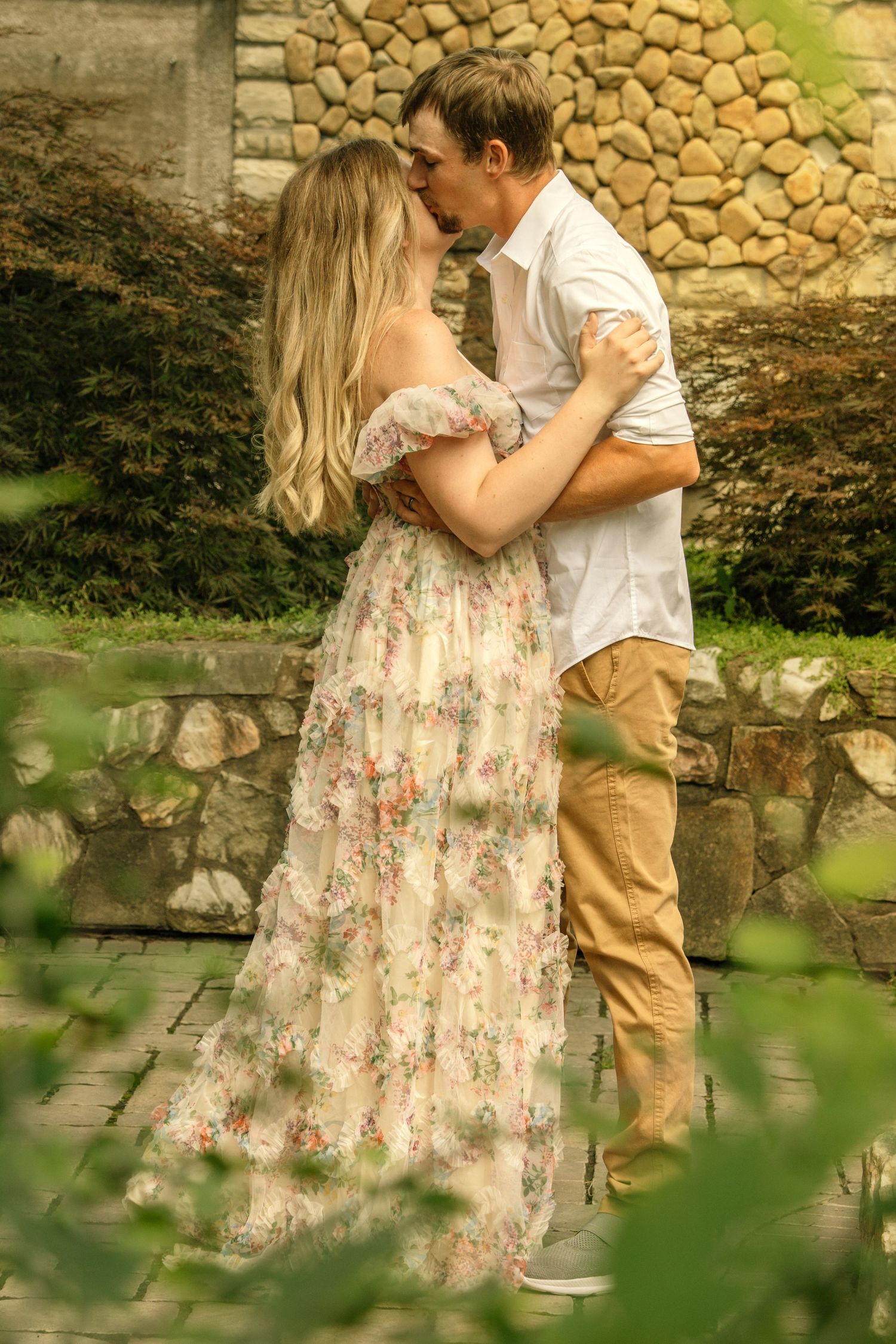Couple sharing a romantic kiss outdoors, woman in floral maxi dress embracing man in white shirt near stone wall.
