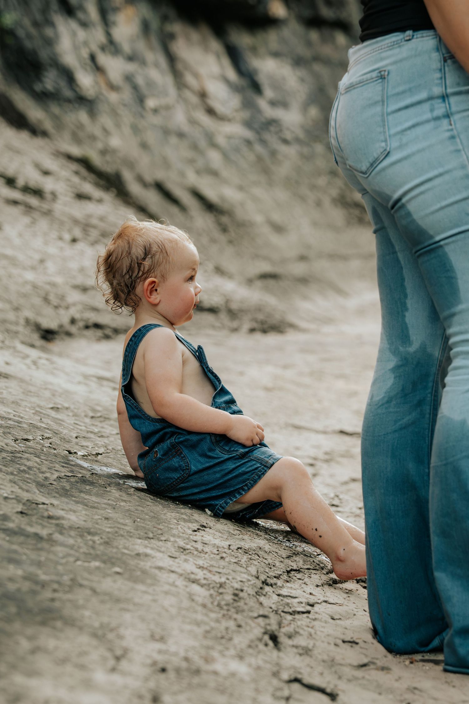 Cute toddler in denim overalls sitting on sandy ground next to mother in blue jeans outdoors.