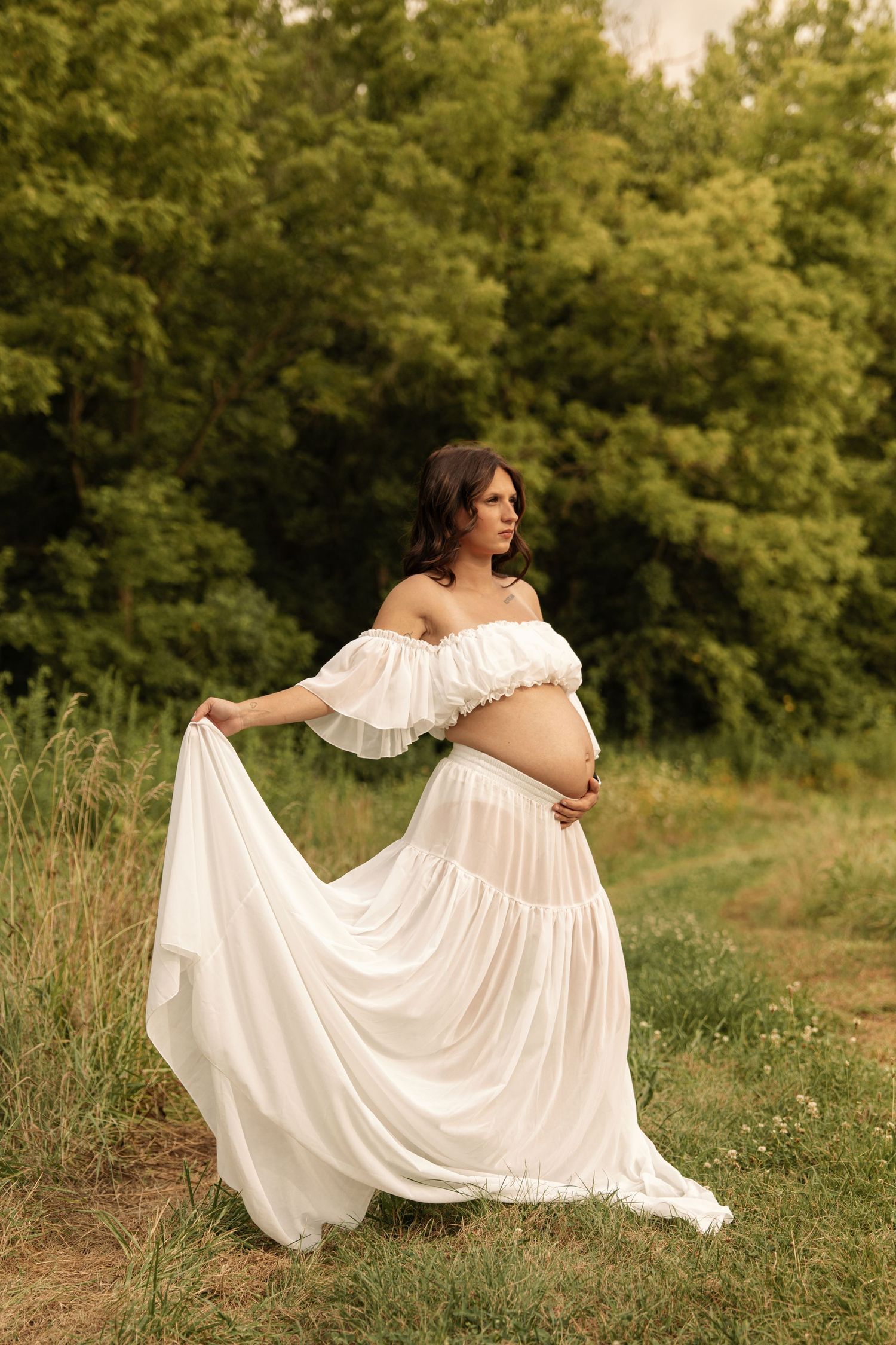 Pregnant woman in a white off-shoulder dress holding her skirt while standing in a green meadow.