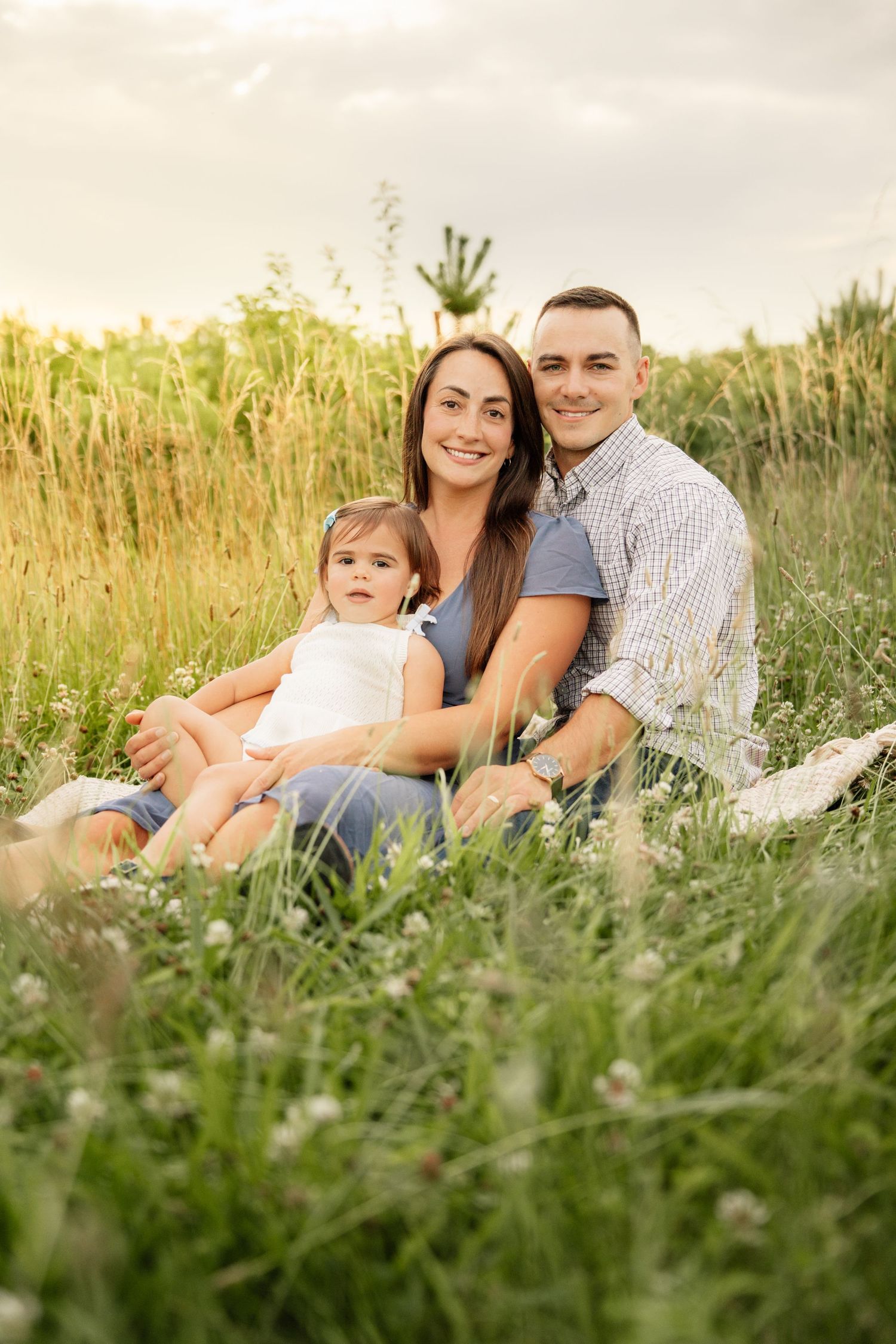 Happy young family with baby girl sitting in green meadow field during golden hour sunset outdoors.
