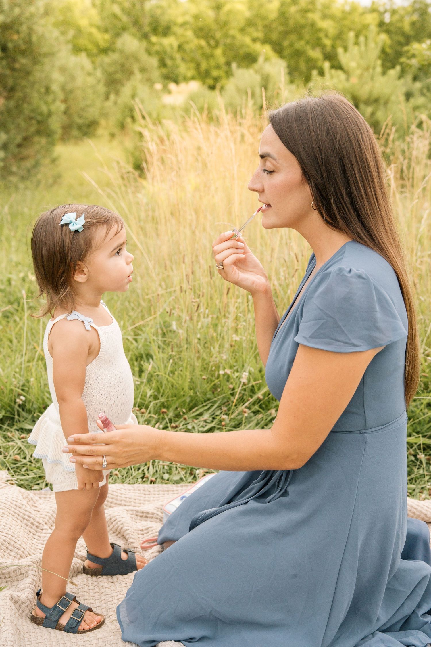 Young mother in blue dress interacts with her toddler daughter on a blanket in a green field.