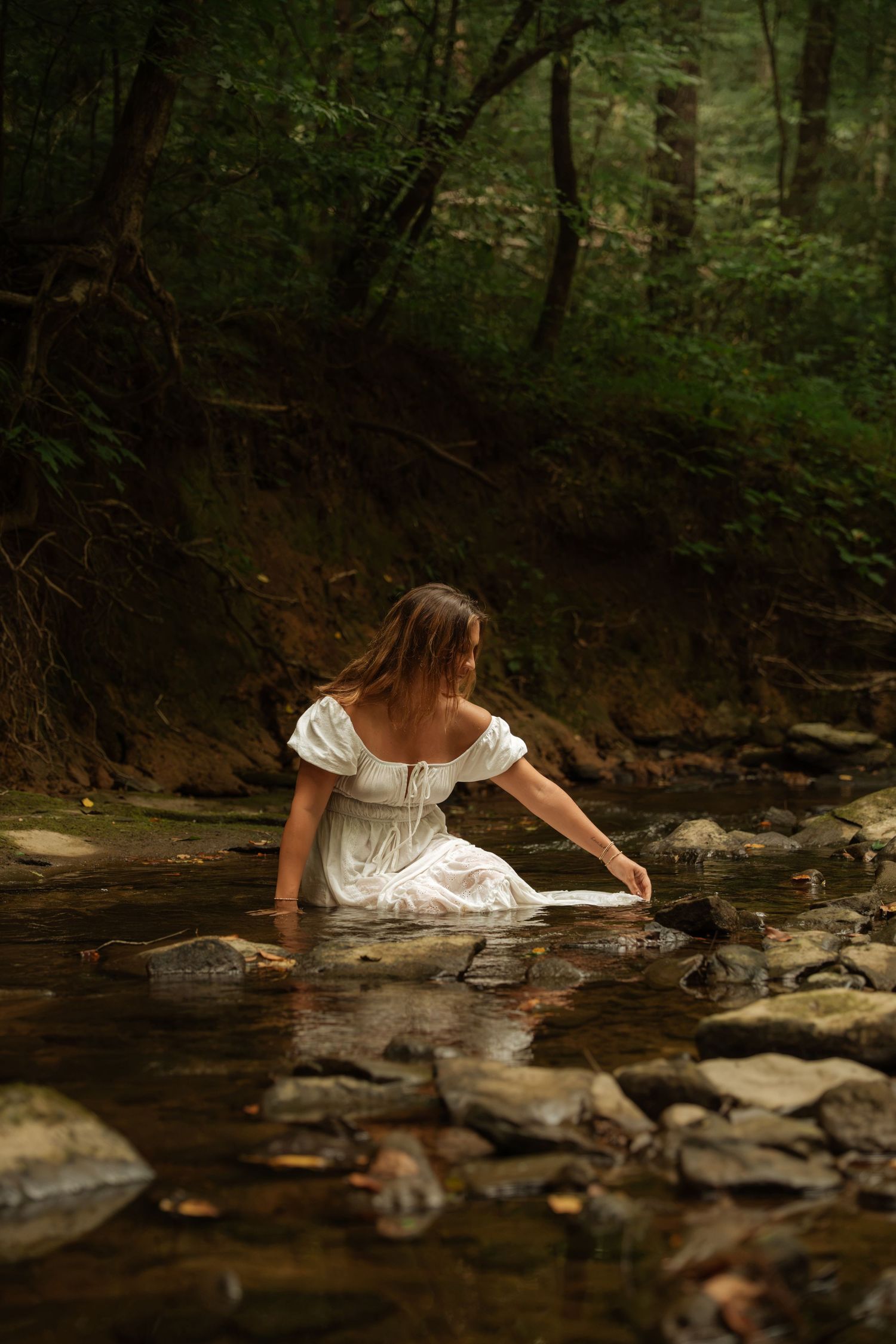 Woman in white dress wading in a rocky forest stream, reaching toward the water.