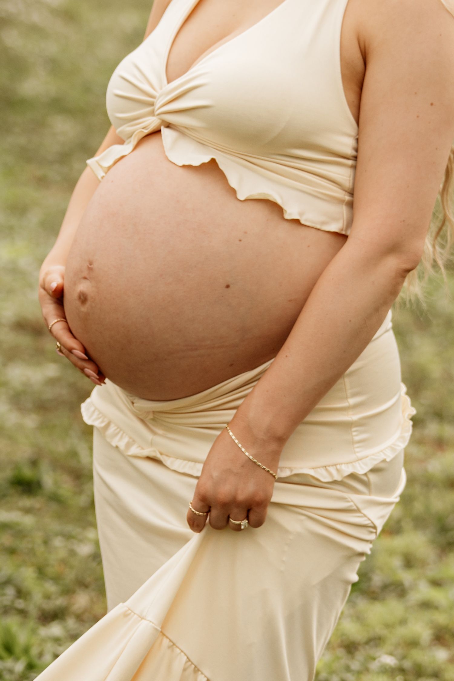Pregnant woman in cream two-piece maternity outfit cradling baby bump outdoors, wearing gold rings and bracelet.