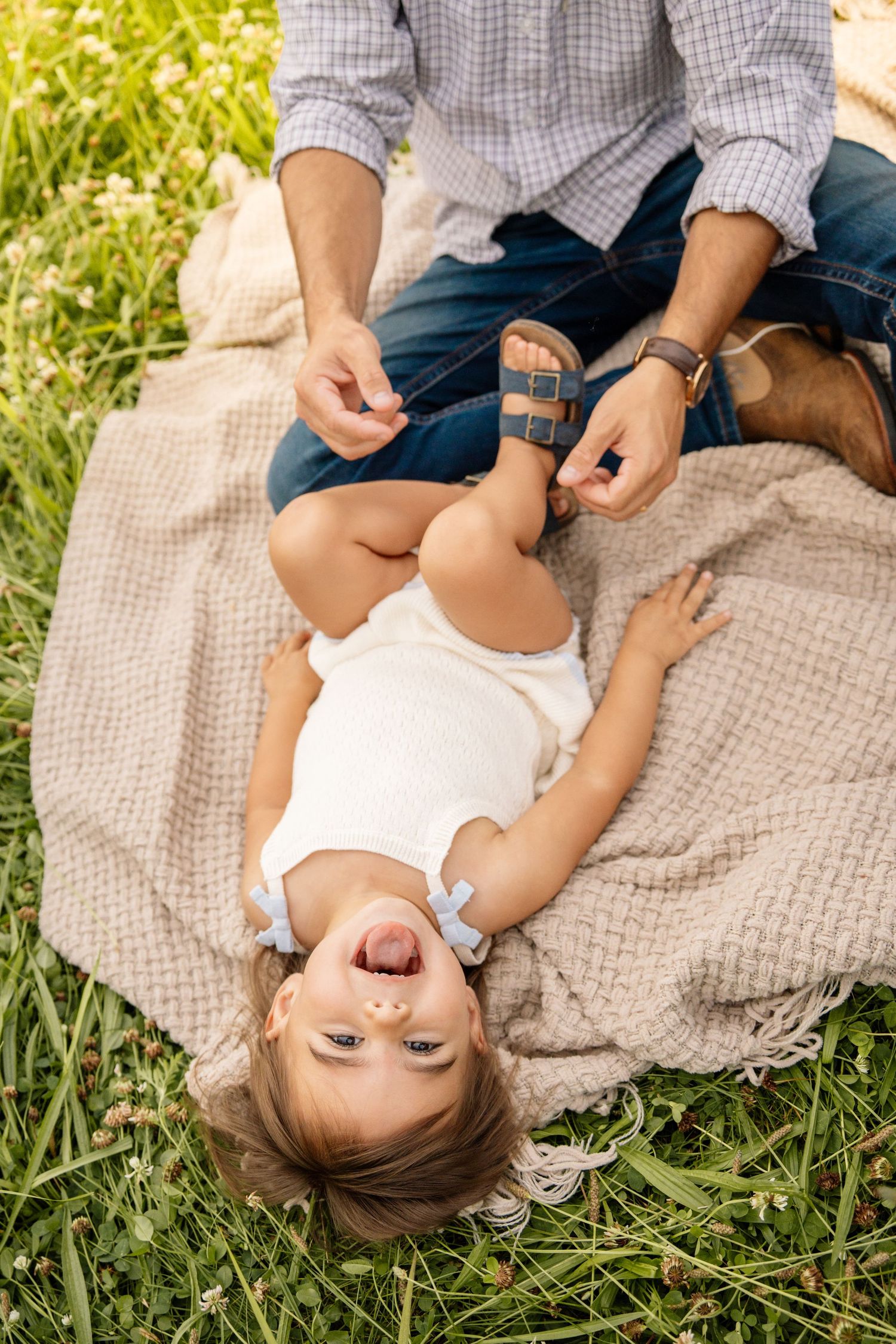 Father playfully holding toddler girl's feet on a blanket in a green grassy field.