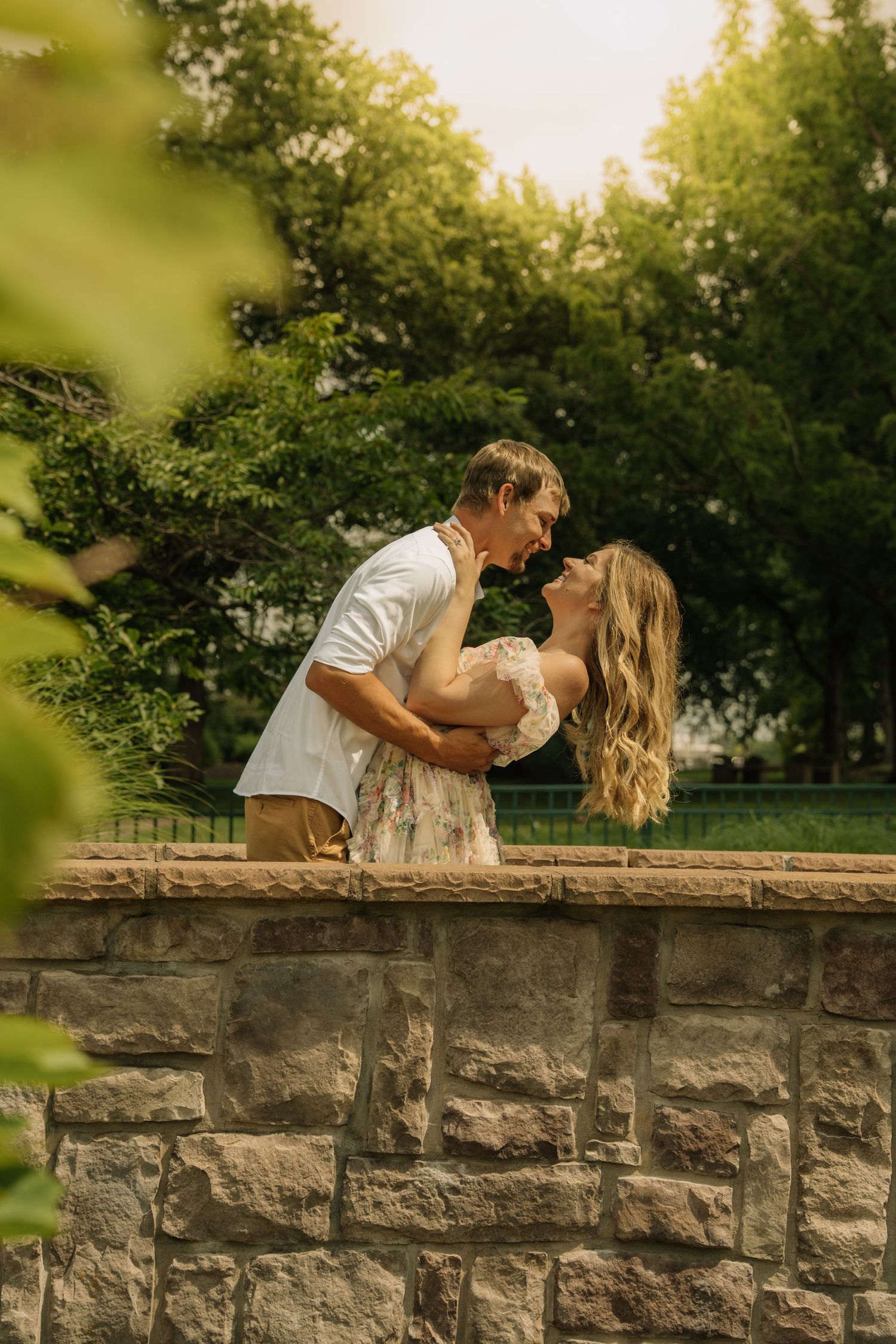 Couple sharing a romantic kiss on a stone wall surrounded by lush green trees in golden hour light.