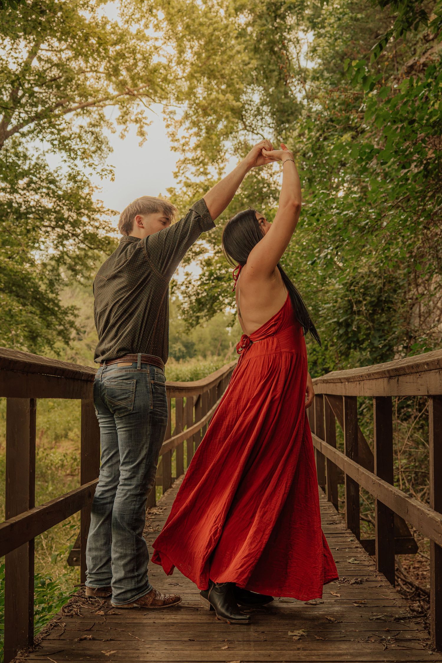 Couple dancing on a wooden bridge surrounded by lush green trees, woman in flowing red backless dress being twirled.