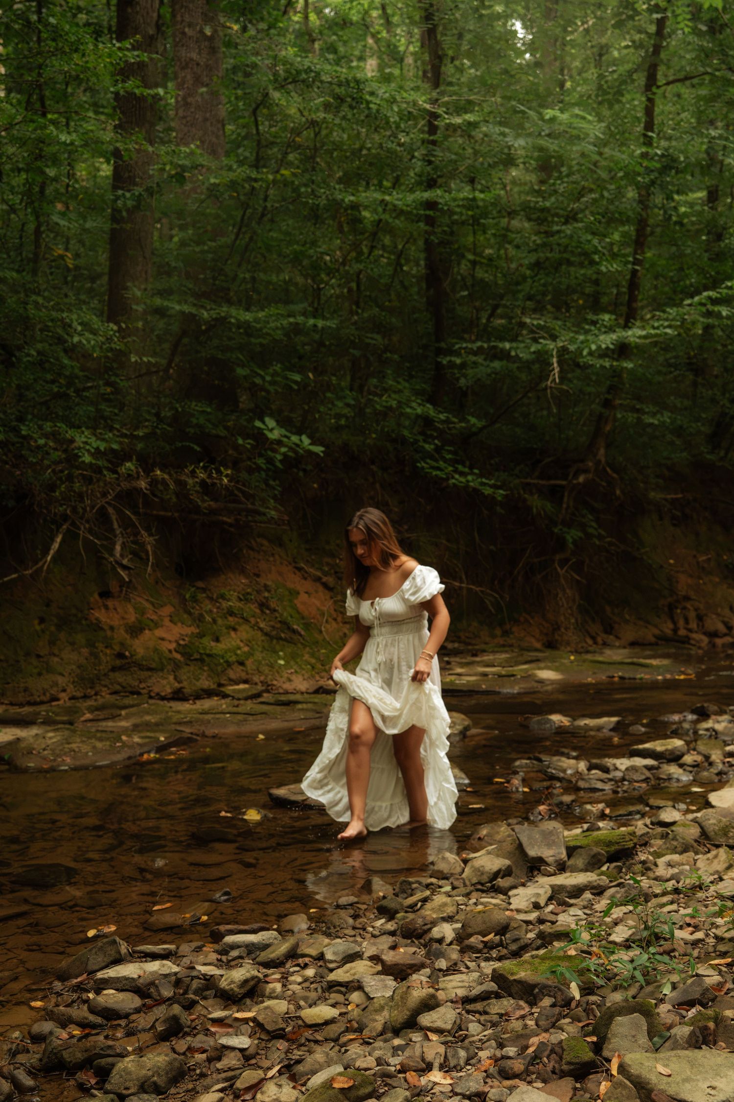 Barefoot woman in white dress walking through a shallow rocky forest stream.