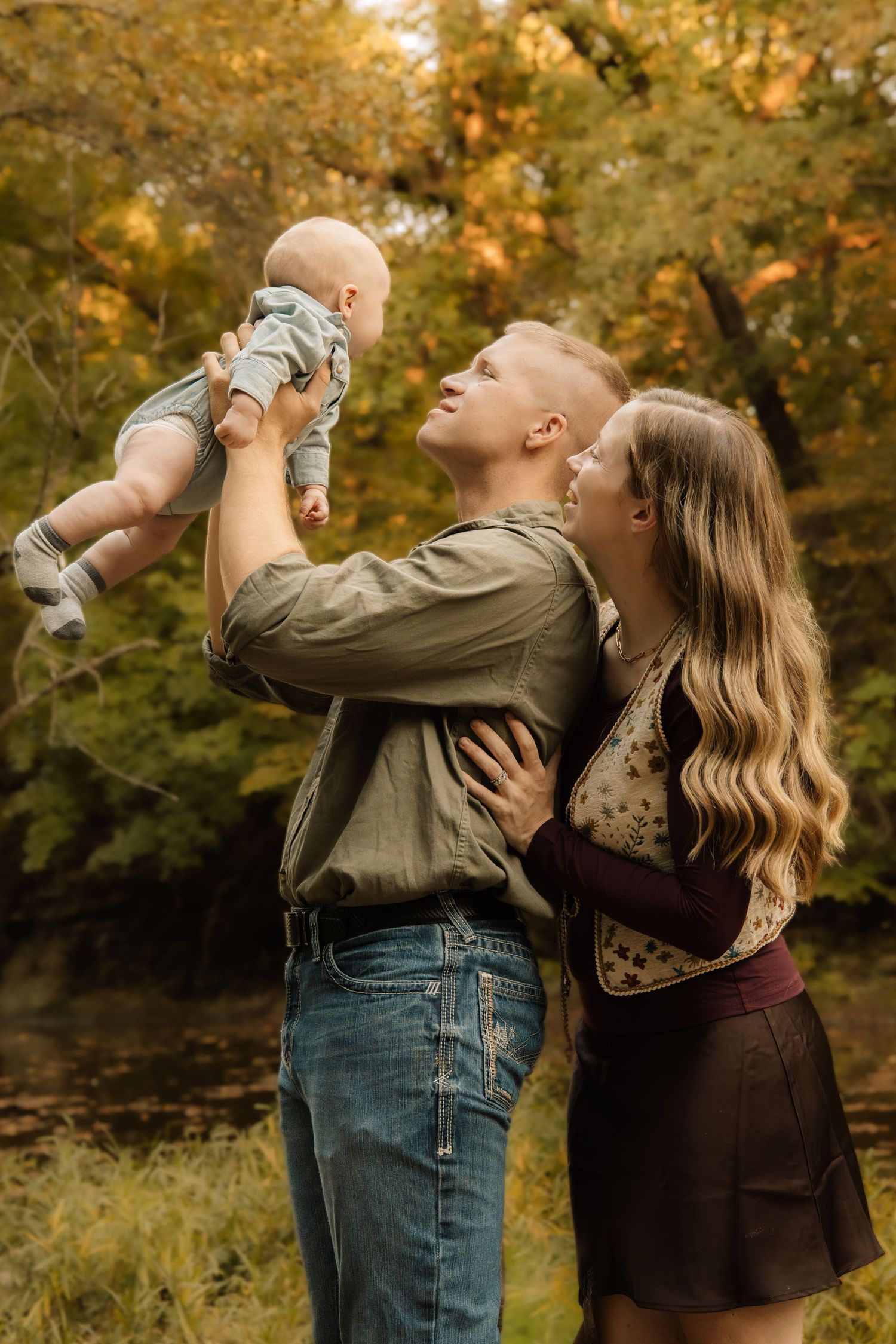Young couple holding baby in autumn forest setting, smiling joyfully during fall family photo session outdoors.