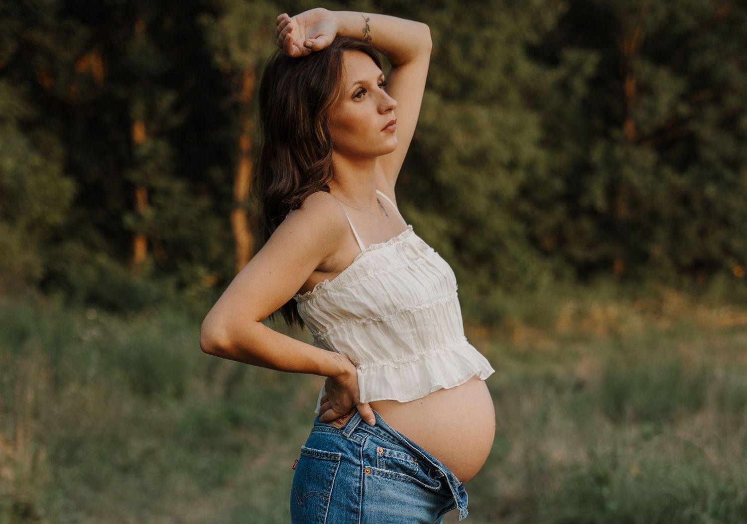 Pregnant woman in white crop top and jeans posing outdoors in a natural green field setting.