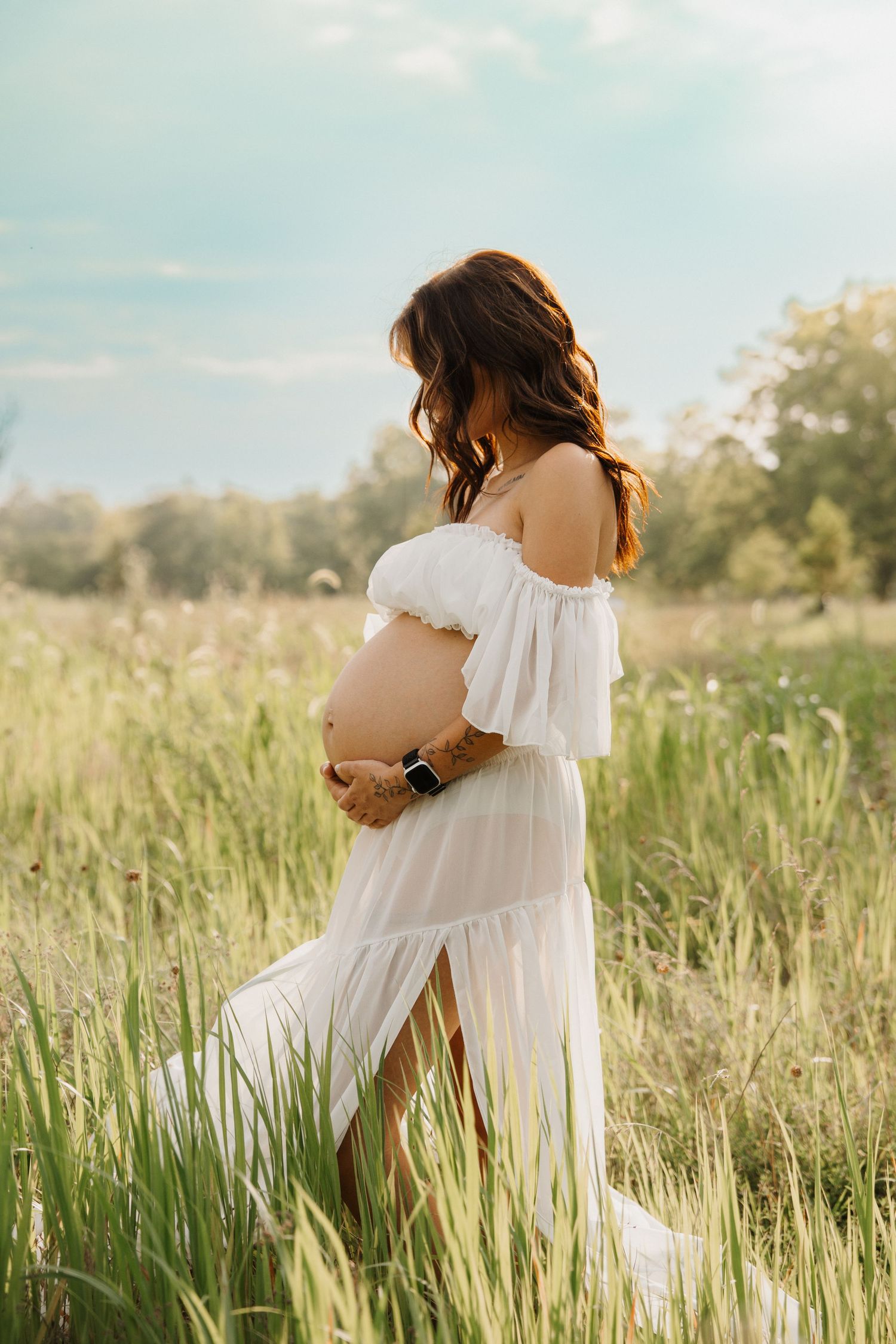 Side profile of pregnant woman in white off-shoulder dress holding baby bump in a golden sunlit field.