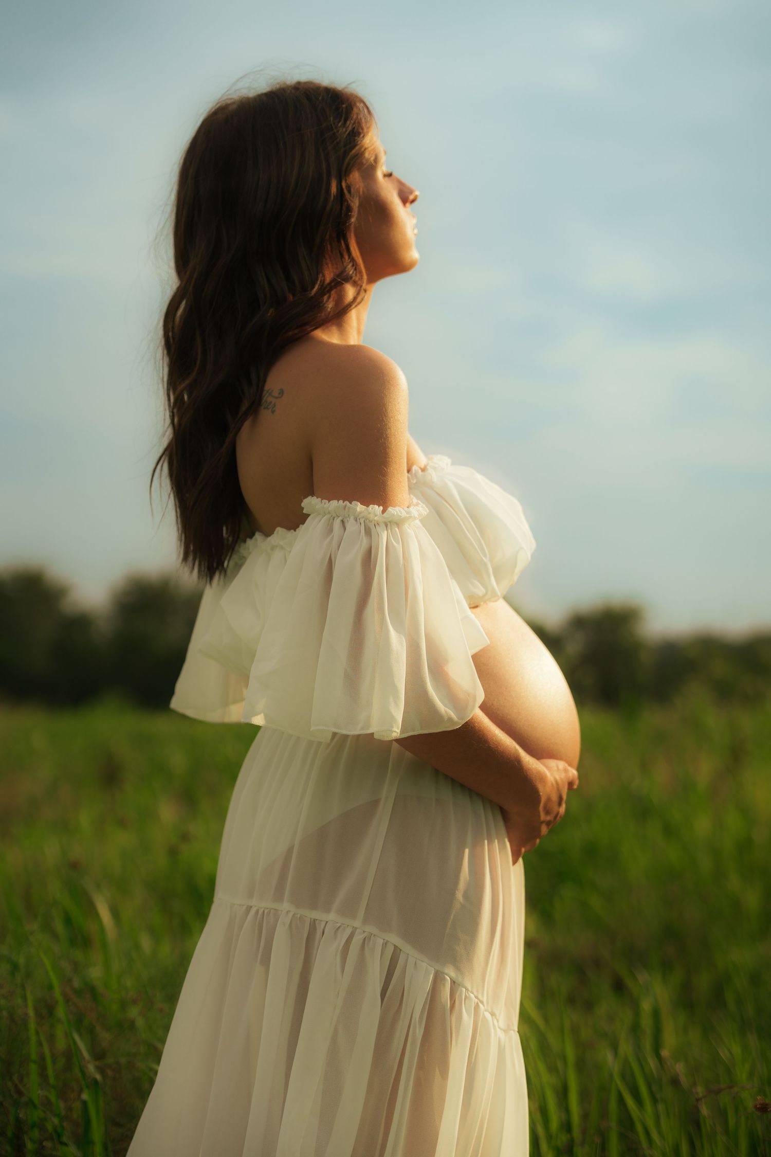 Pregnant woman in sheer white off-shoulder dress cradles baby bump while gazing upward in green field.