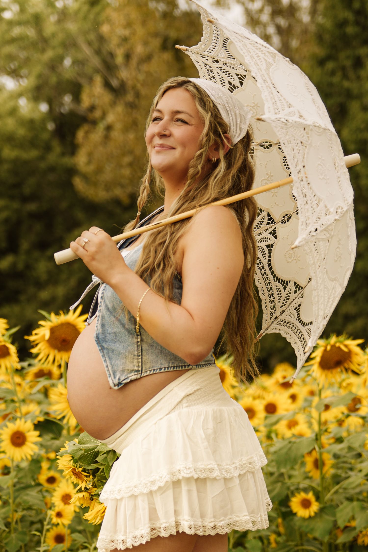 Pregnant woman in sunflower field holding lace parasol, wearing denim crop top and white skirt, smiling outdoors.