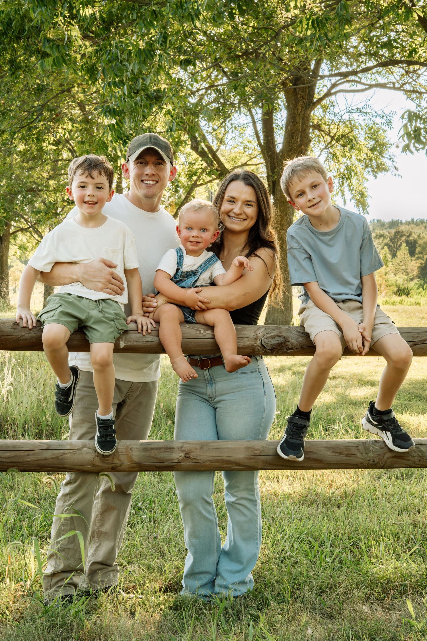 Happy family of five posing outdoors by a wooden fence, surrounded by lush green trees on a sunny day.