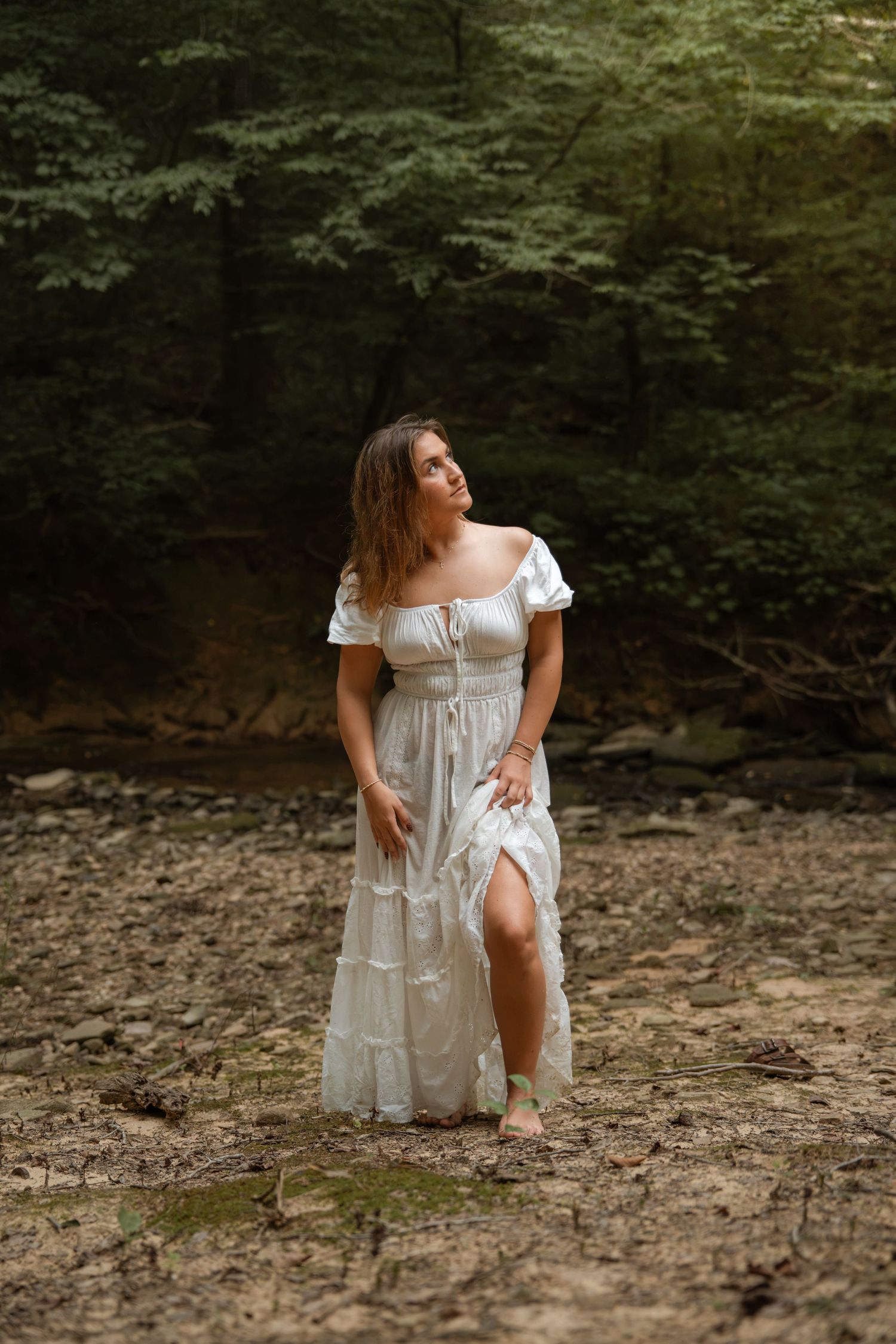 Woman in white off-shoulder dress walks barefoot on dry rocky forest creek bed.
