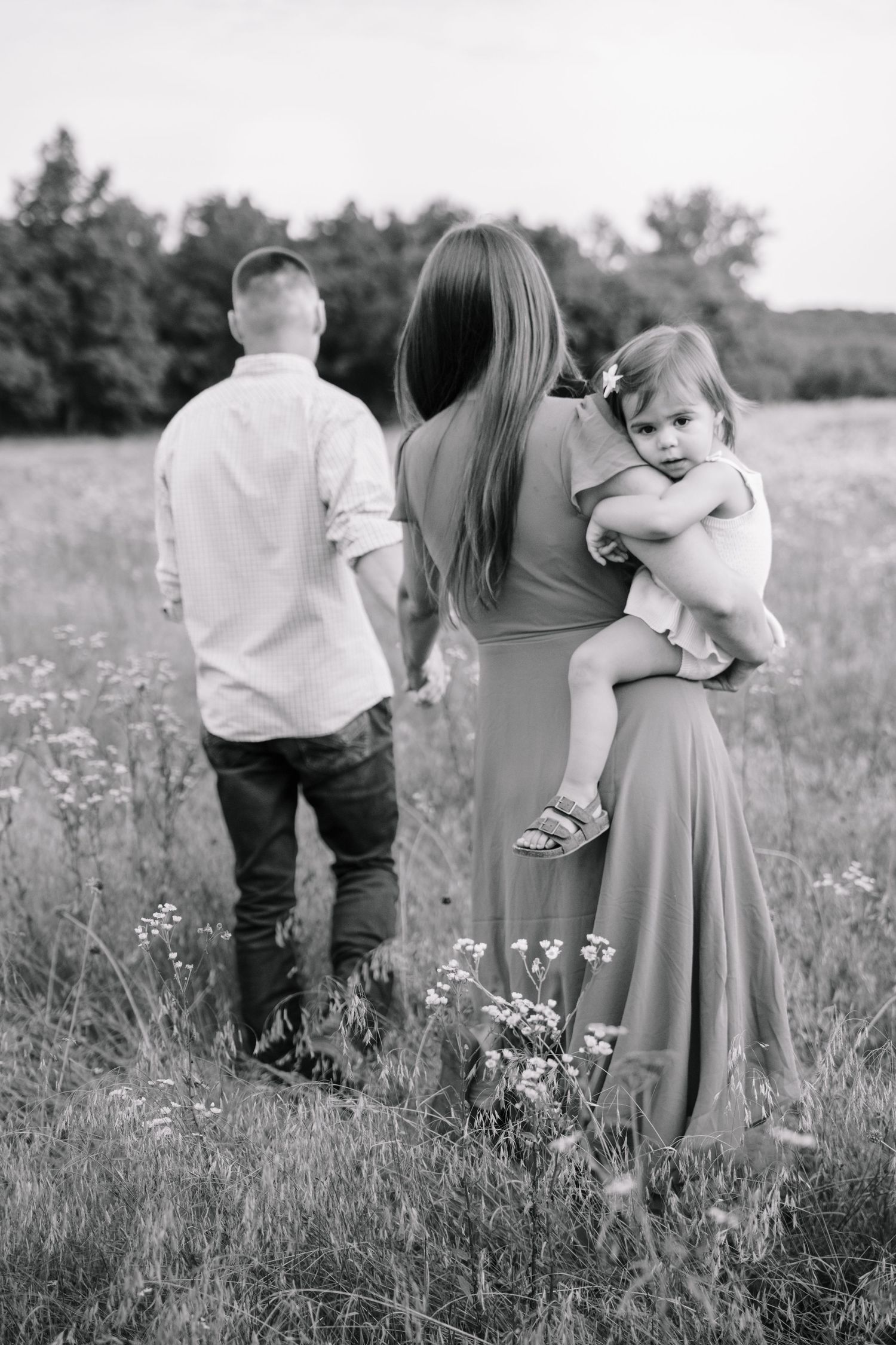 Black and white photo of a family in a field, mother holding toddler while father walks ahead.