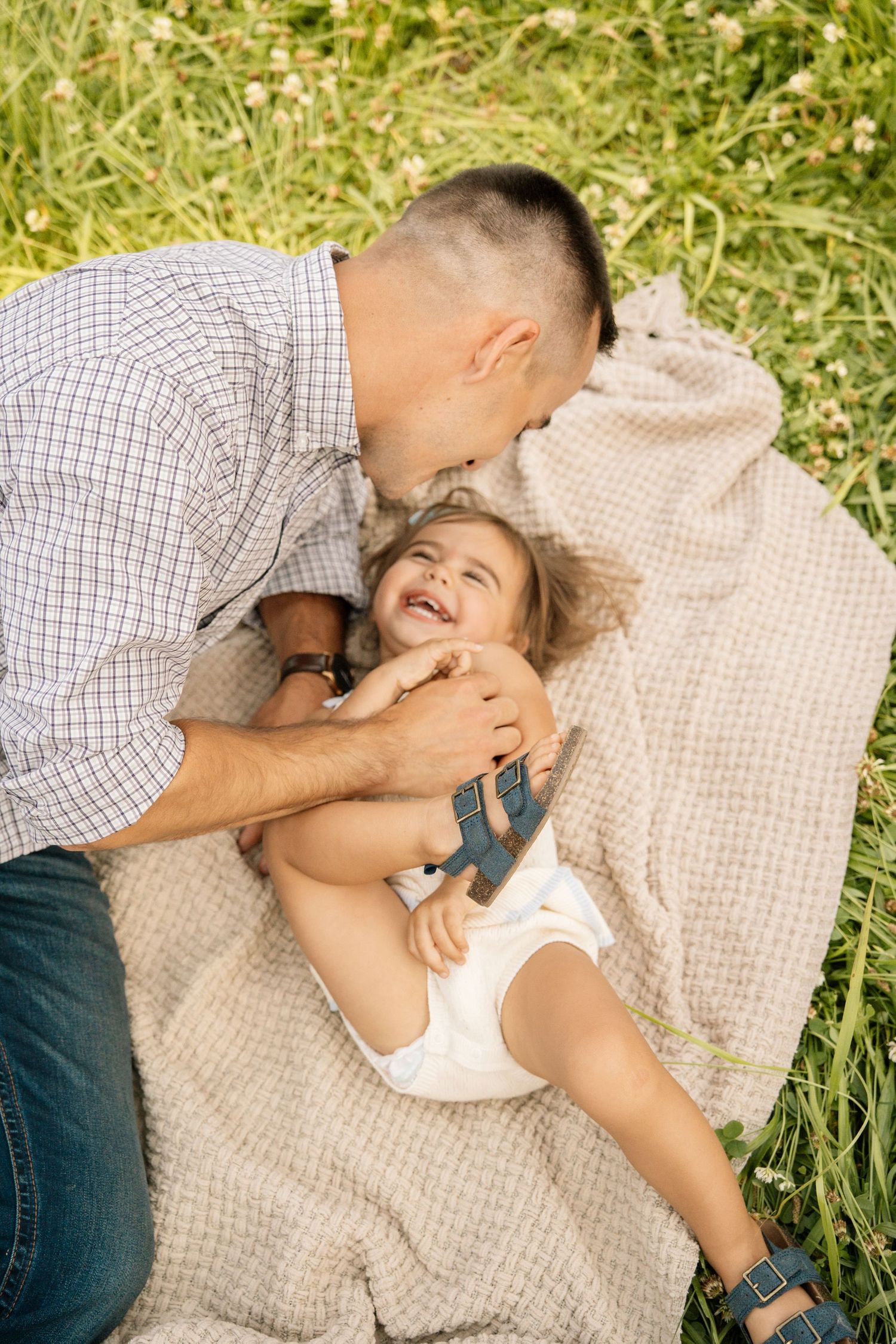 Father playfully tickles laughing toddler daughter lying on blanket in green grass.