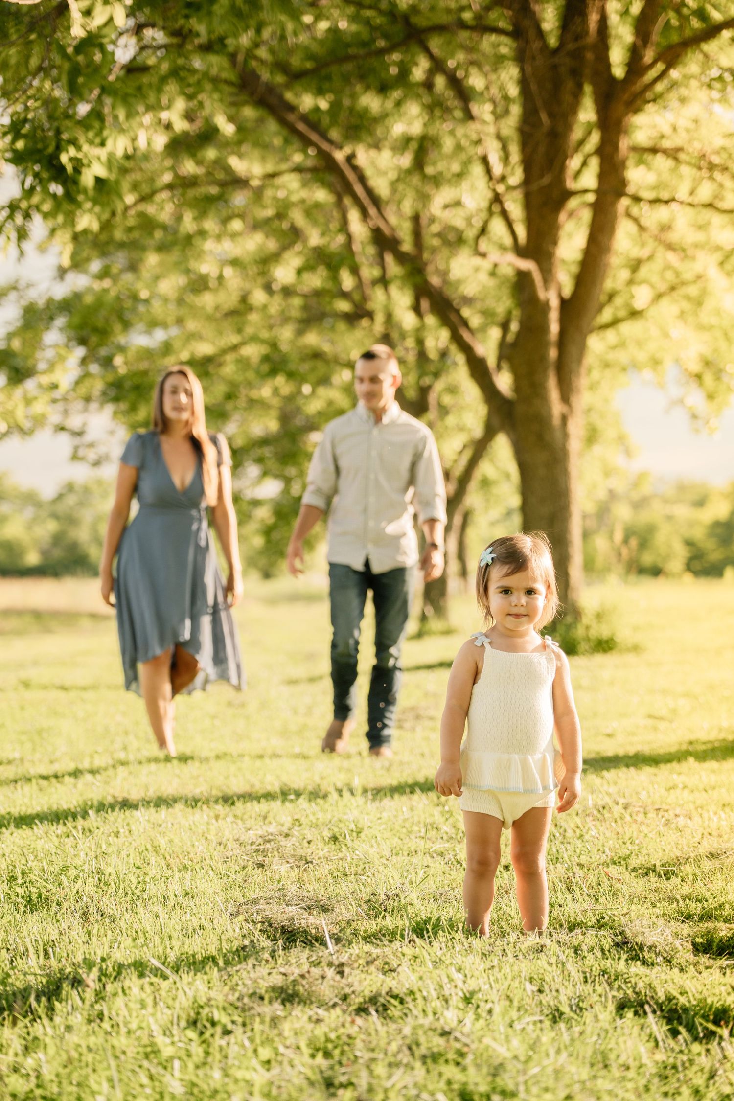A toddler girl walks toward the camera in a sunny park while her parents stroll hand-in-hand behind her.