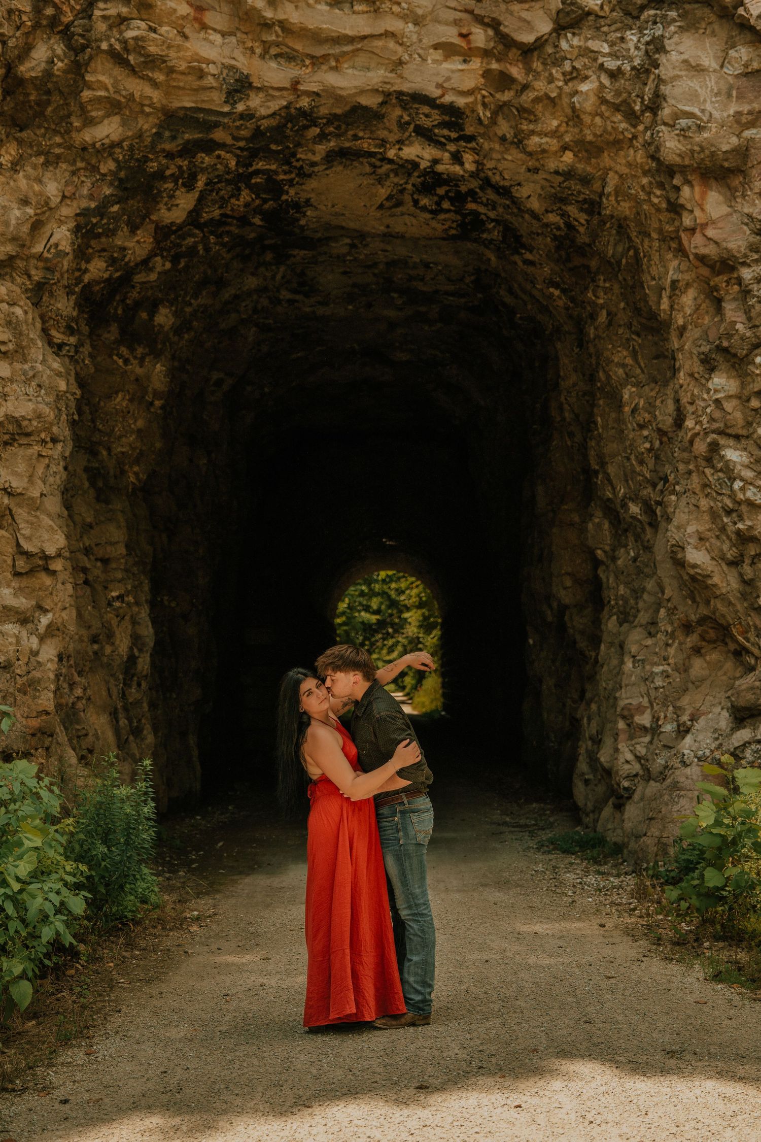 Couple embracing romantically in a rustic rock tunnel, woman in red dress, lush greenery visible through the arched stone passage.